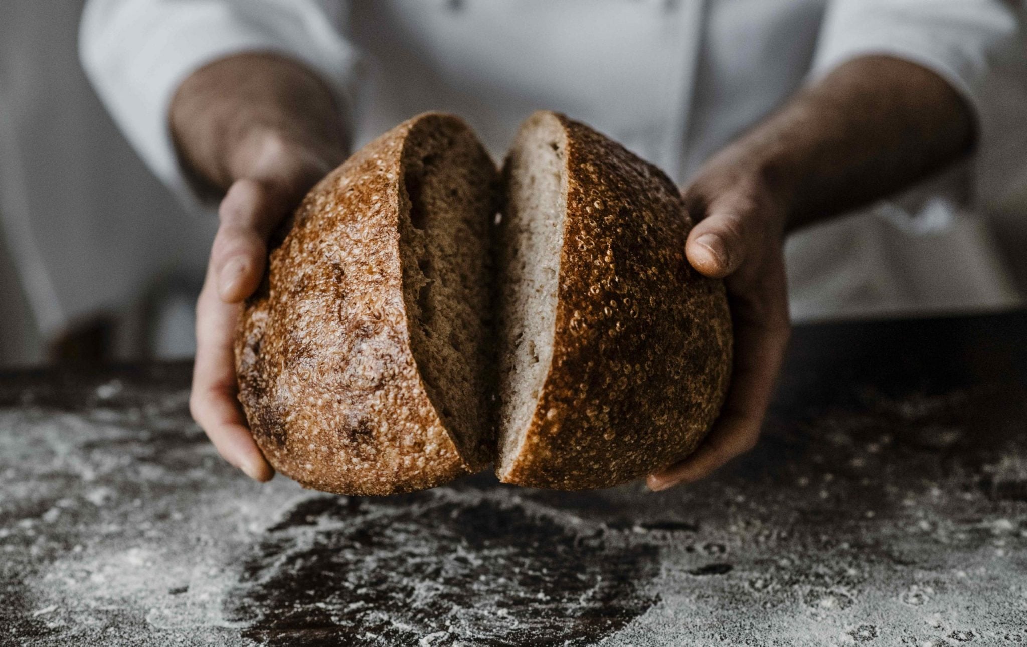 Casa di Langa, Europe — Baker's hands breaking open artisanal sourdough loaf with flour-dusted crust on marble surface.