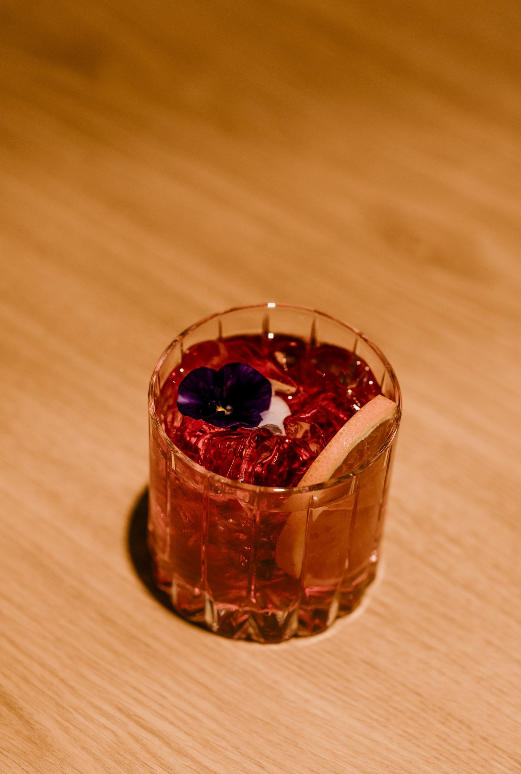 Sorì Cocktail Bar, Europe — Amber cocktail with crushed ice and dark pansy flower garnish, warm wooden surface and soft natural light.