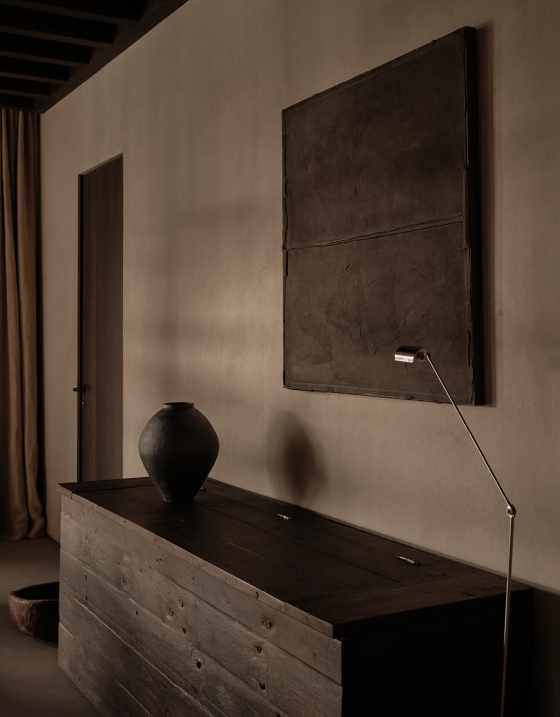En Kyanó, Europe — Minimalist bedroom with wall-mounted television, dark wood dresser, black ceramic vessel, and sculptural pendant lighting in neutral tones.