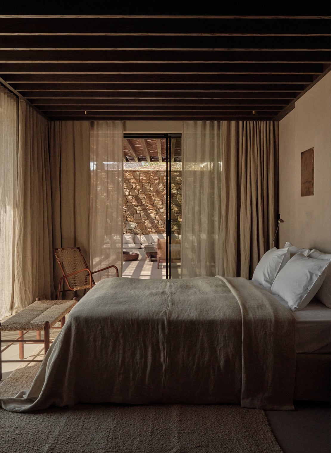 En Kyanó, Europe — Minimalist bedroom with exposed timber ceiling, linen bedding, sheer curtains framing terracotta landscape view, woven chair, warm natural light.