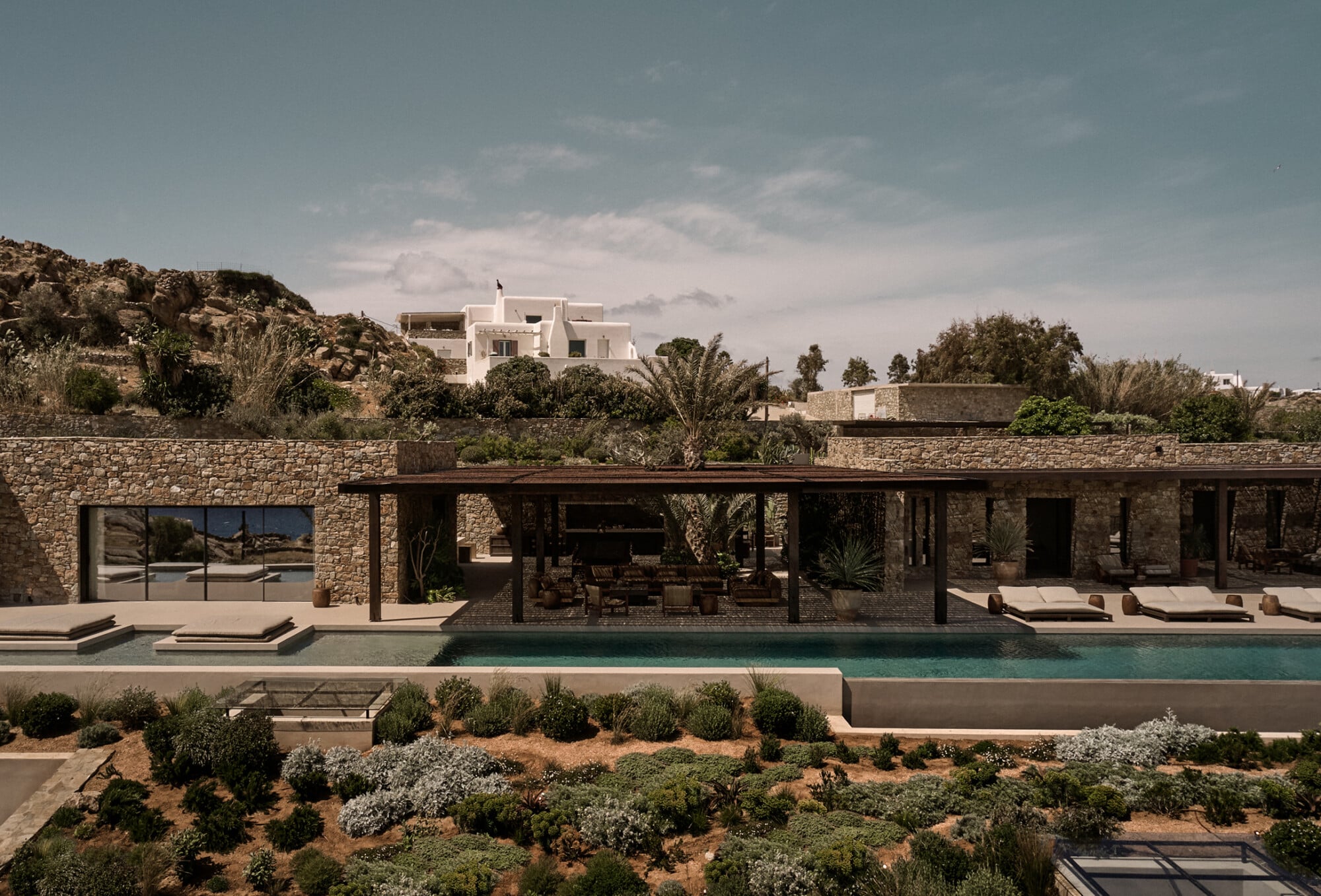 En Kyanó, Europe — Modernist stone villa with turquoise pool, wooden pergola, Mediterranean landscaping, and rocky hillside setting in warm afternoon light.