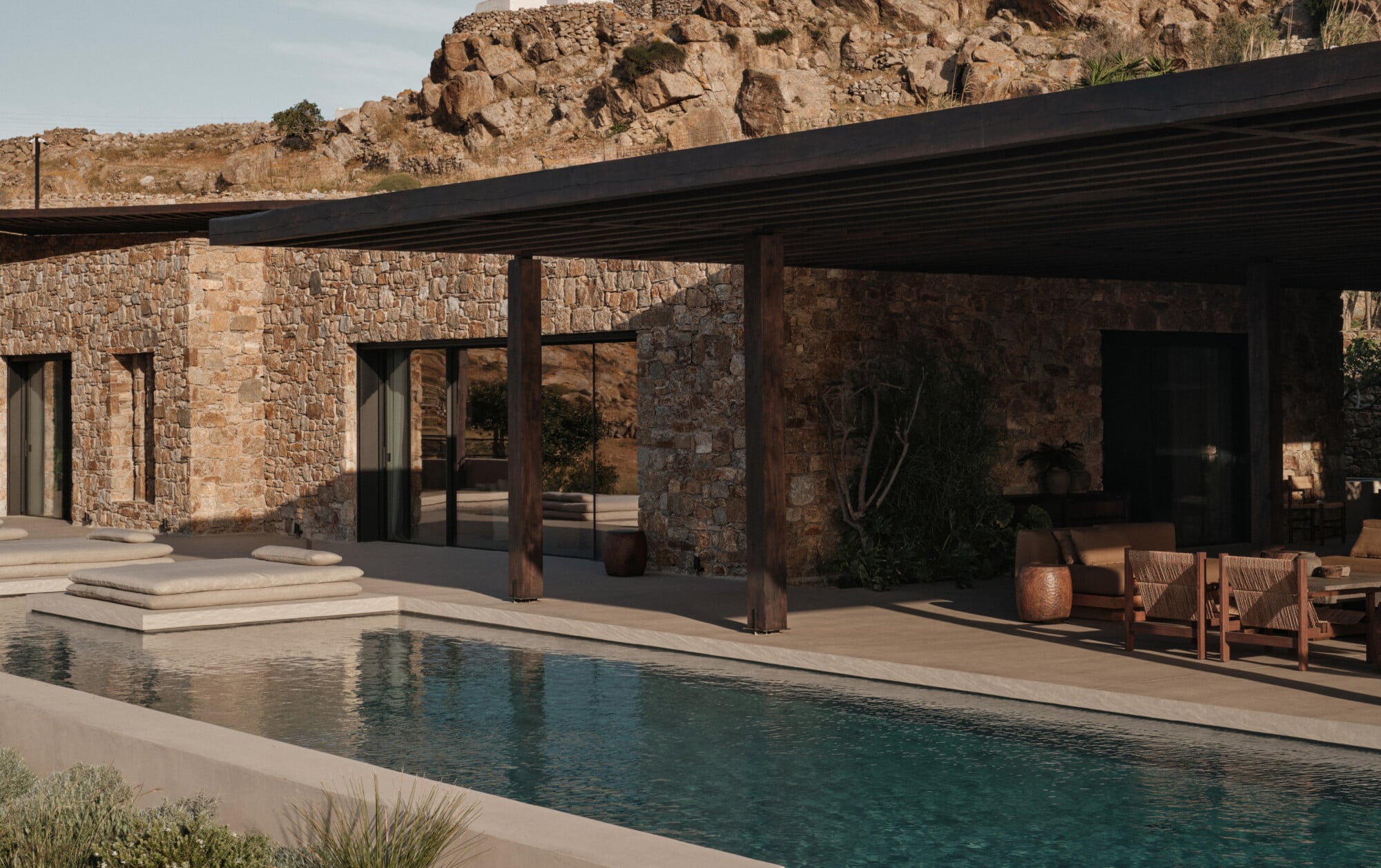 En Kyanó, Europe — Modernist stone villa with black steel pergola overlooking lap pool, terracotta pottery, and rocky hillside backdrop in warm Mediterranean light.