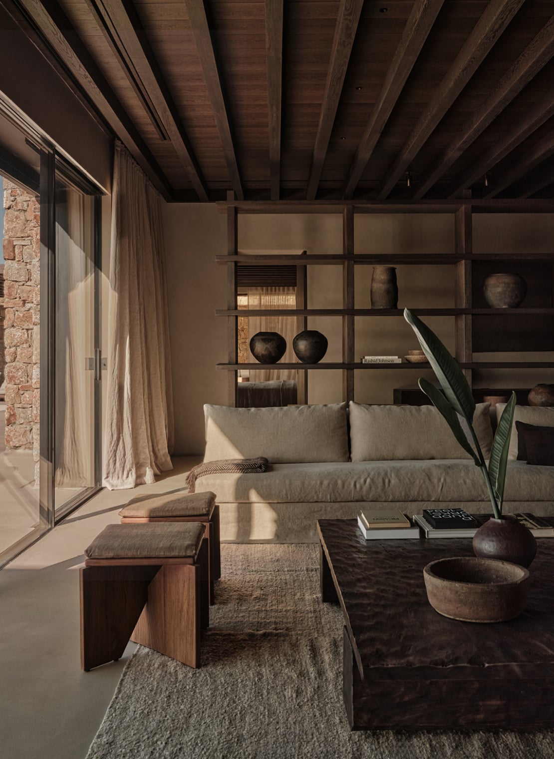 En Kyanó, Europe — Minimalist living room with cream sofa, dark wood ceiling beams, gridded shelving wall, natural light, sculptural ceramics, and stone fireplace.
