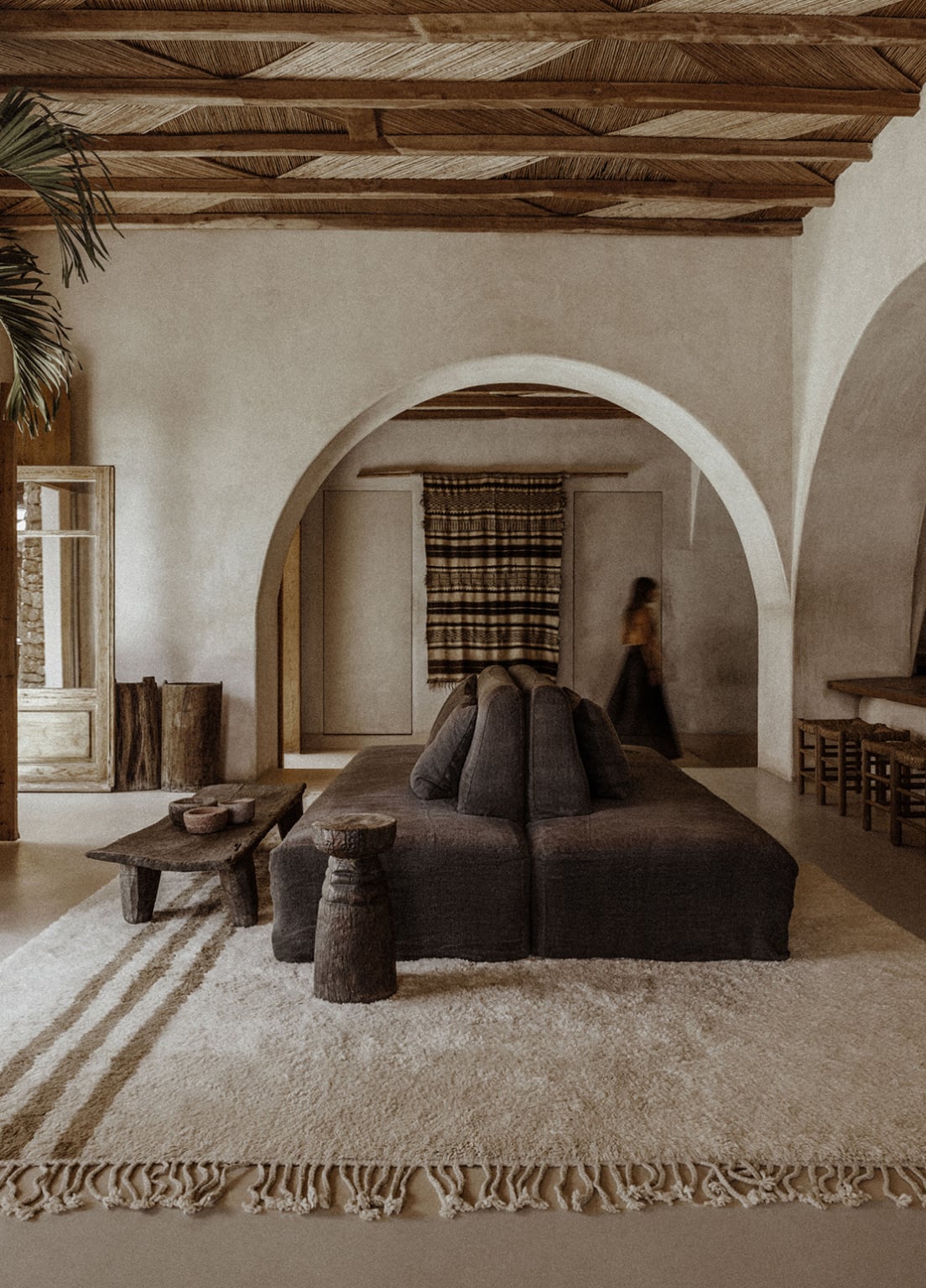 En Kyanó, Europe — Minimalist bedroom with arched doorways, exposed wooden beams, charcoal linen bed, woven textiles, and natural light casting shadows on whitewashed walls.