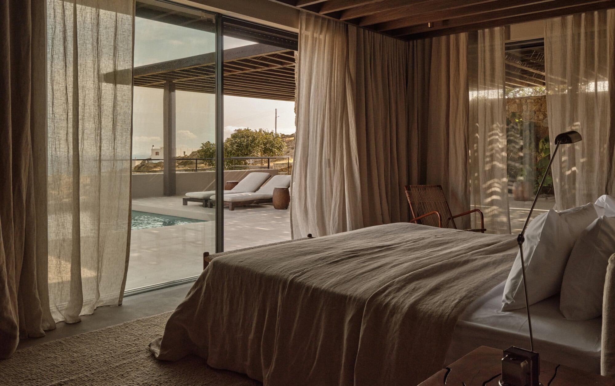 En Kyanó, Europe — Bedroom with linen curtains, wooden ceiling, stone terrace, lounge chairs, and pool overlooking Mediterranean landscape.