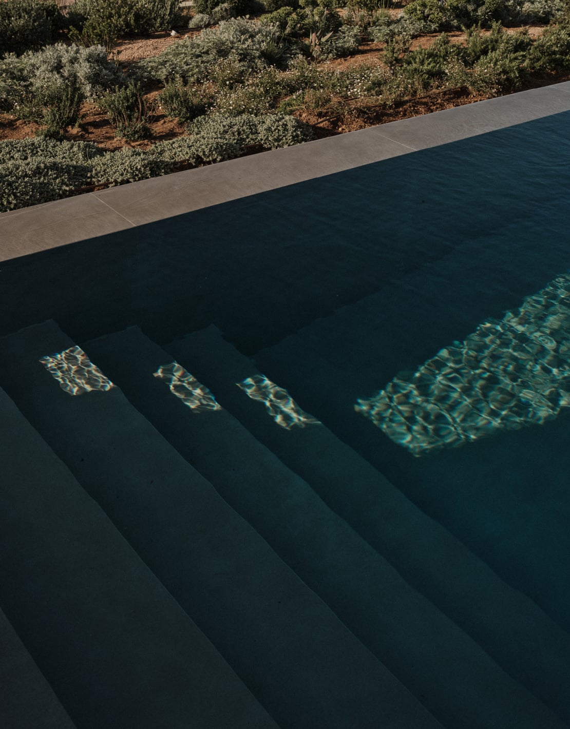 En Kyanó, Europe — Dark-tiled infinity pool with stepped entry and golden light reflections, overlooking olive groves and terracotta landscape.