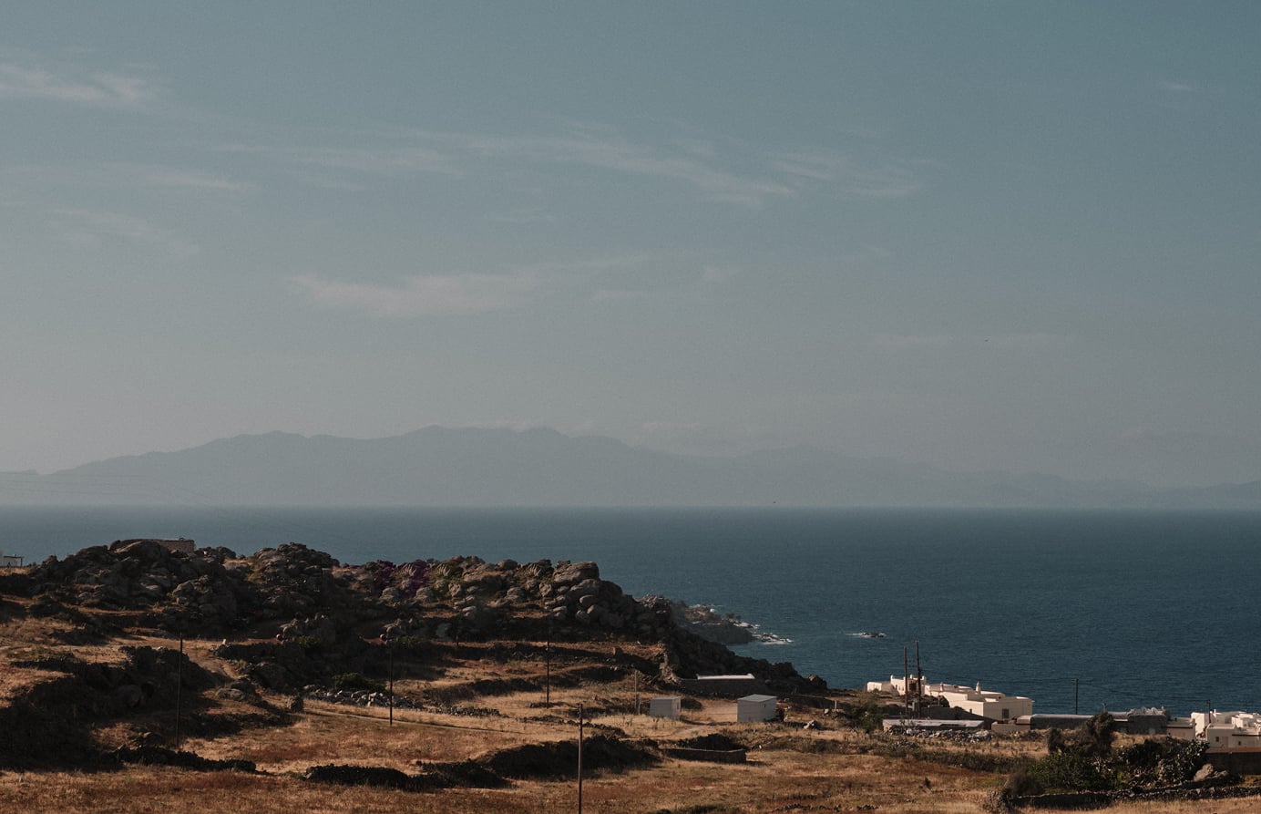 En Kyanó, Europe — Coastal hillside with whitewashed buildings overlooking turquoise sea, rocky outcrops, and distant mountains under hazy sky