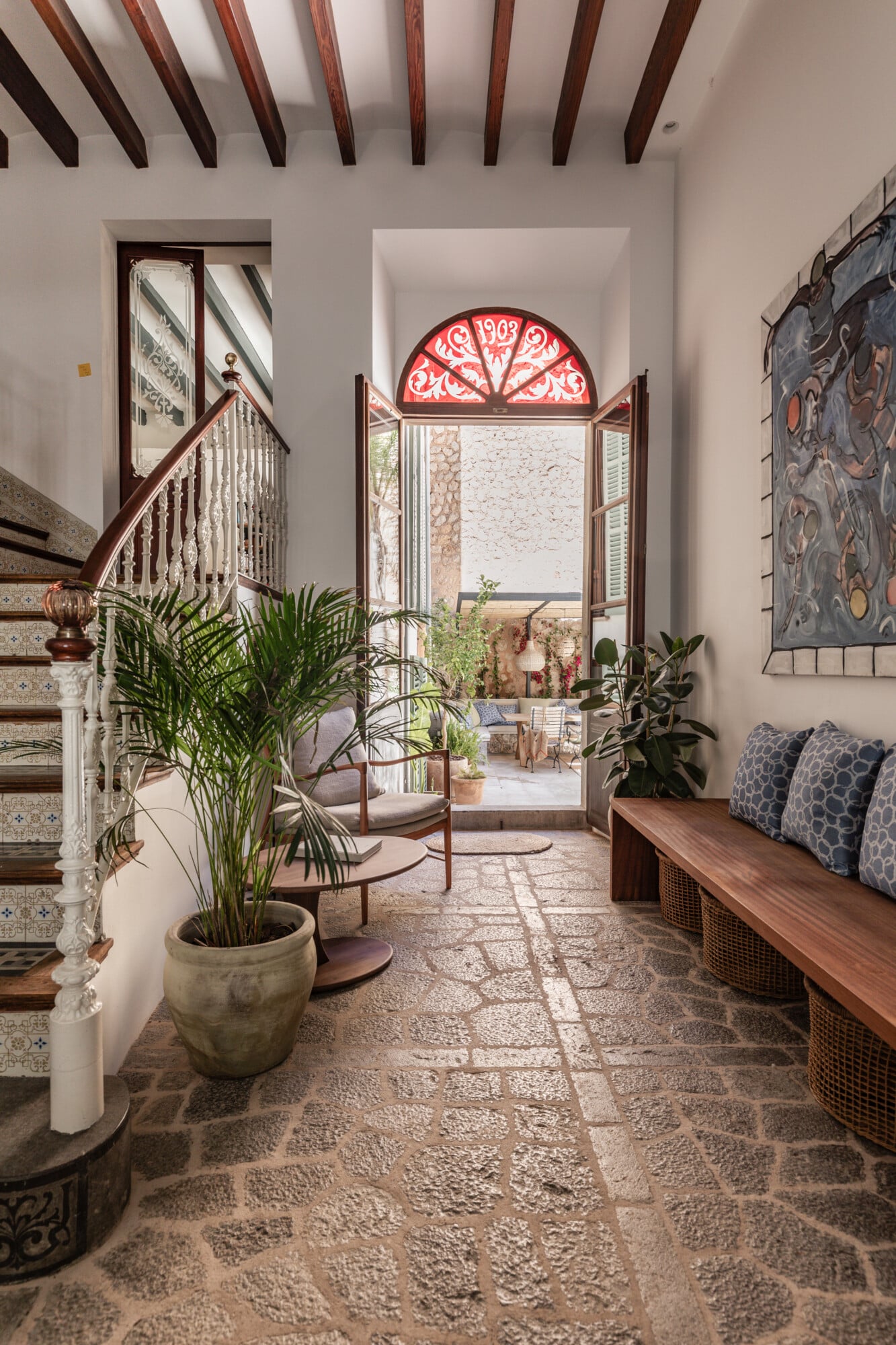 Mon Cor, Balearic Islands, Spain — Bright hallway with exposed wooden beams, Moorish arched window, terracotta tile floor, potted palms, wooden bench with blue pillows, ornamental wall art.