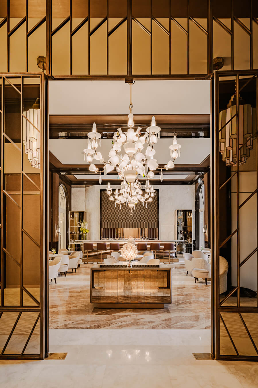 Carlton Cannes, Cannes, France — Sunlit lobby with brass-framed doors, sculptural white chandelier, marble floors, and modernist furnishings in warm neutrals.
