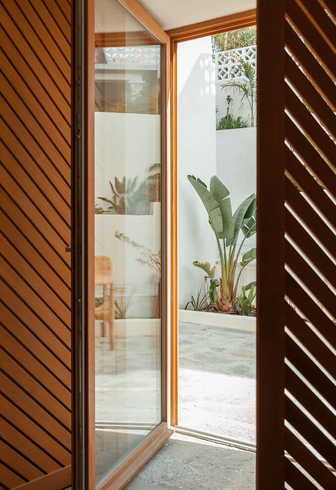Casa Santa Teresa, Europe — Open wooden-frame glass door with slatted wood panels revealing minimalist courtyard with banana plants and white walls, warm natural light and shadows.