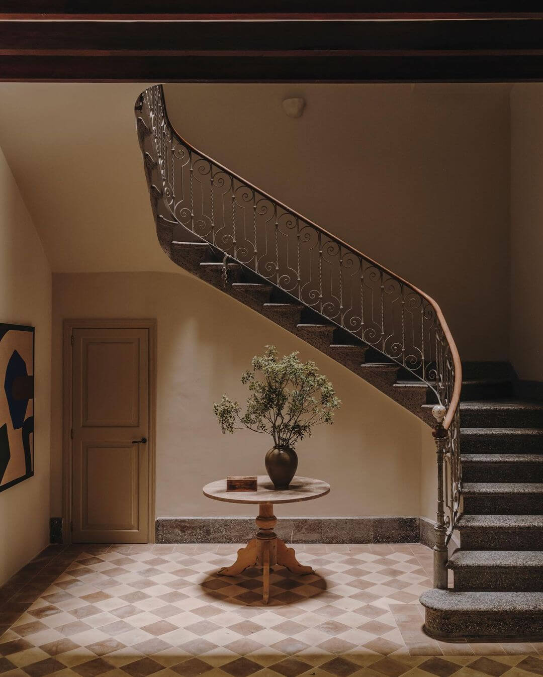 Santa Teresa 34, Balearic Islands, Spain — Curved wrought-iron staircase with ornamental railings descending beside marble pedestal table with dried botanicals in bronze vase, cream walls, geometric floor tiles.