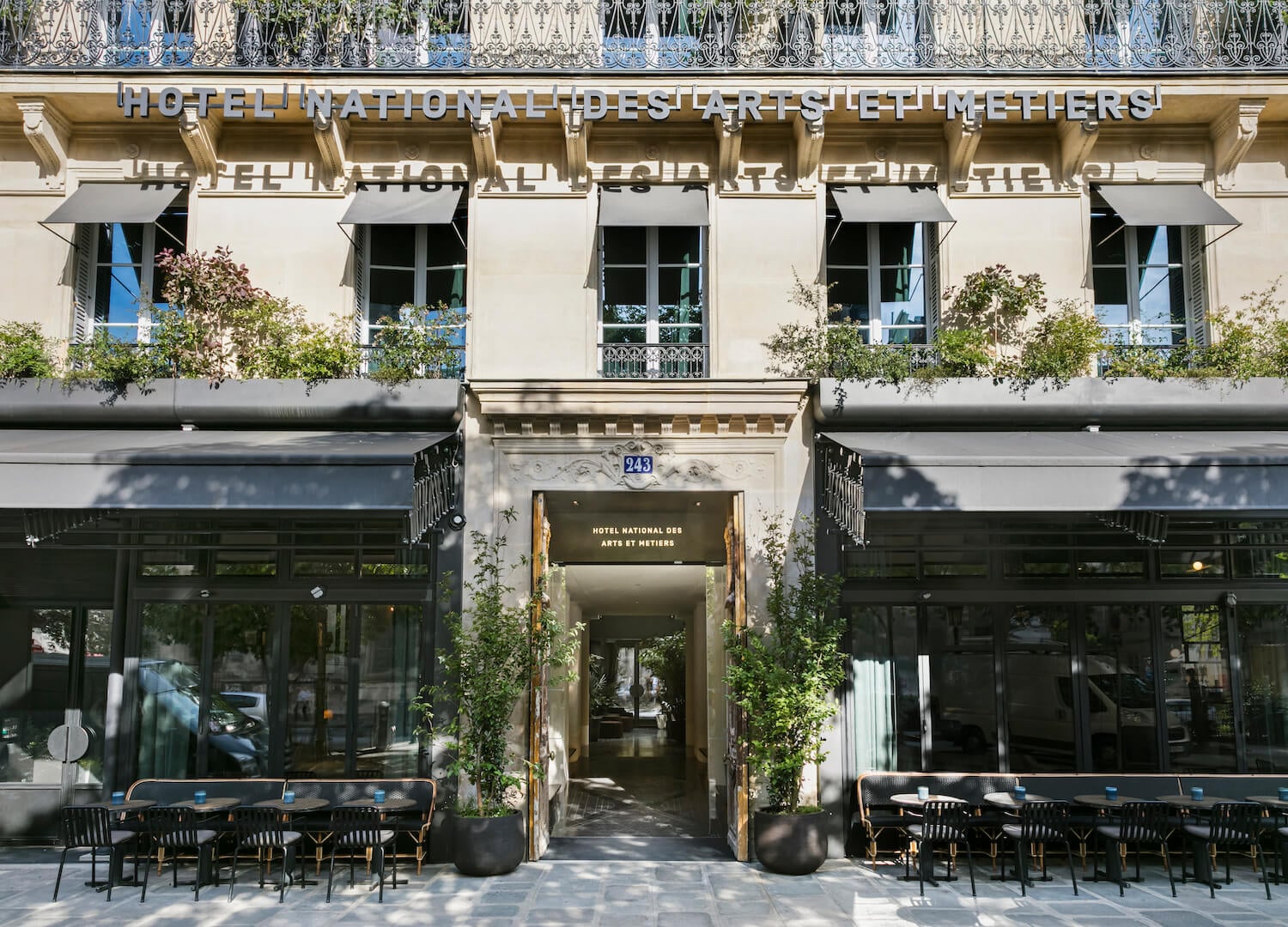 Hôtel National Des Arts, Paris, France — Neoclassical façade with black awnings, potted greenery, and outdoor seating beneath ornate stonework and wrought-iron balconies.