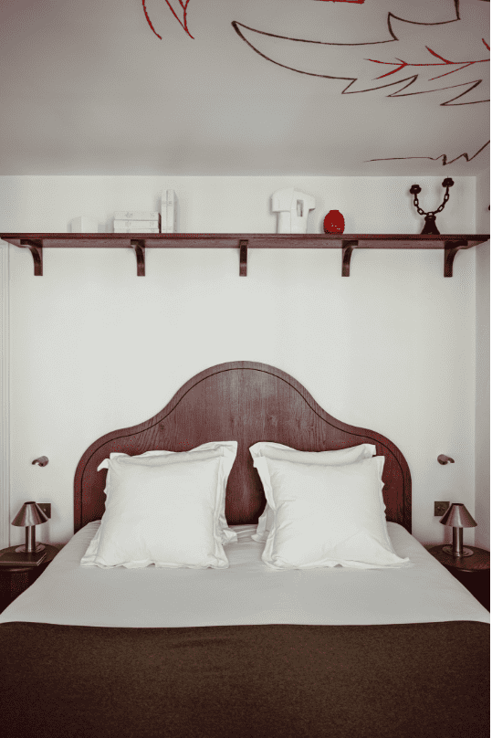 Académies et des Arts, Europe — Burgundy wood bed headboard with white linens, floating shelf, minimal nightstands, and abstract red wall art in contemporary bedroom interior.