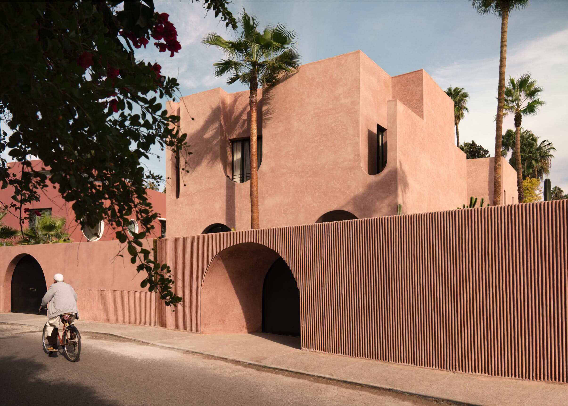 Maison Brummell Majorelle, Africa — Terracotta-hued modernist facade with arched openings, ribbed wall detailing, palm trees, and cyclist in morning light.
