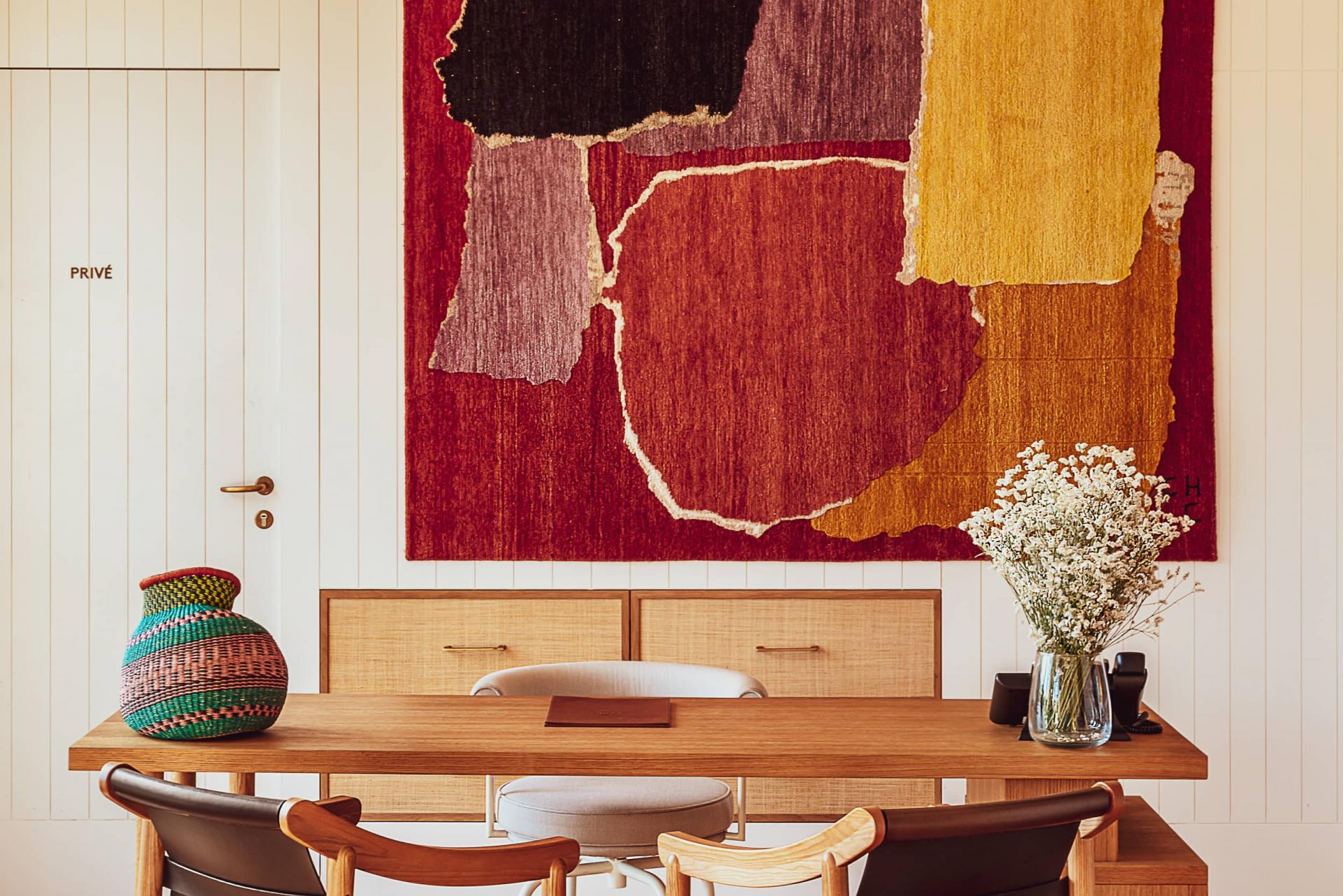 Hôtel Lou Pinet, Europe — Minimalist office with warm wood desk, cream linen chairs, woven basket, dried flowers, and abstract red-gold textile map on white shiplap wall.