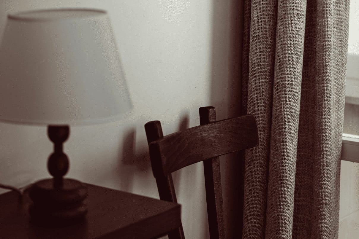 Académies et des Arts, Europe — Dark wooden chair beside cream linen curtains and table lamp in minimalist hotel room with soft natural light.