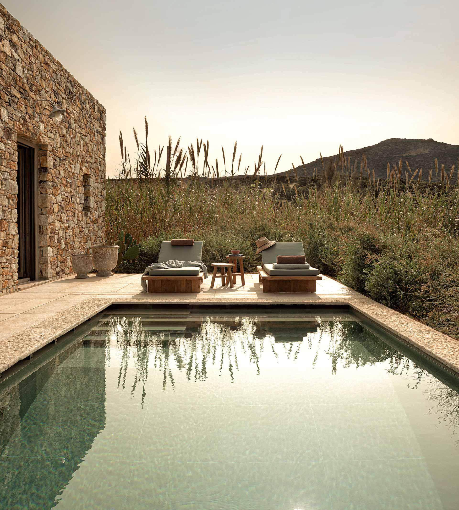 The Rooster, Europe — Limestone terrace with reflecting pool, wooden loungers, stone cottage, and wild grasses under golden hour light.