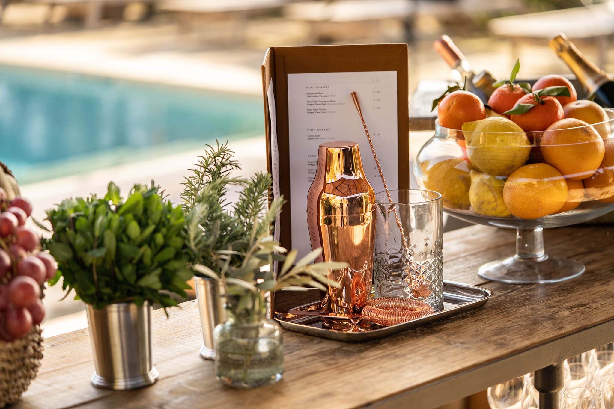 Finca Serena, Balearic Islands — Poolside dining setup with copper cocktail shaker, fresh citrus fruits, potted succulents, and wine menu in afternoon sunlight.