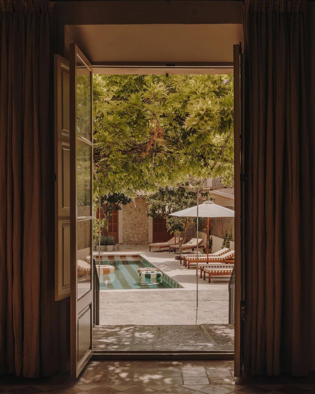 Santa Teresa 34, Balearic Islands, Spain — Open wooden doors framing courtyard with thermal pool, striped loungers, shade umbrella, and lush vine-covered walls in warm Mediterranean light.