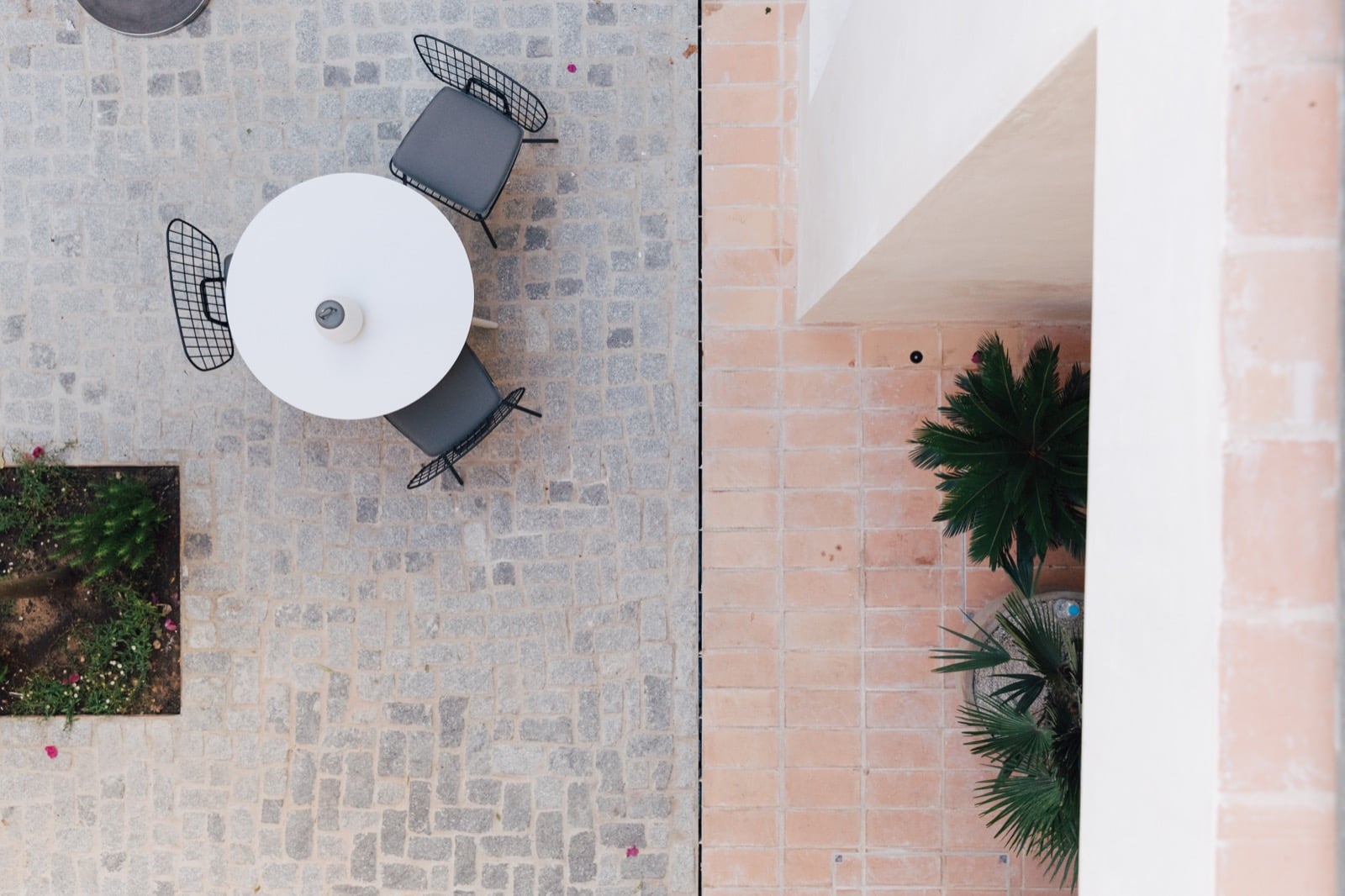 Plácido y Grata, Europe — Overhead view of circular white table with black chairs on stone-tiled courtyard, palm plants and brick architecture visible.