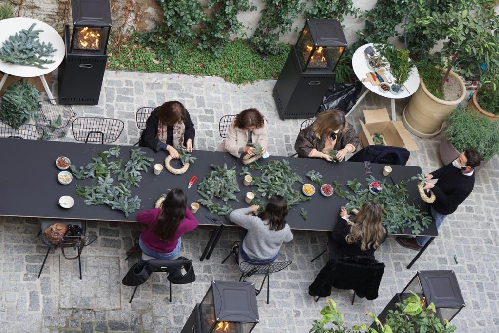 Local Workshops, Europe — Overhead view of guests seated at black table with fresh herbs and ingredients, ivy-covered courtyard with fire heaters and stone paving.