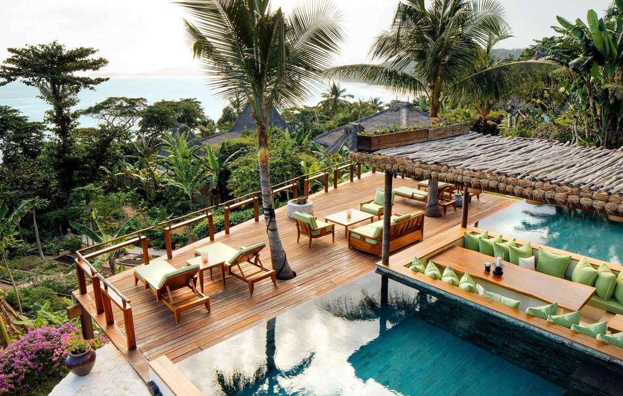 NIHI Sumba, Indonesia — Teak terrace with lounge seating overlooking turquoise infinity pool, tropical gardens, and ocean view at luxury resort villa.