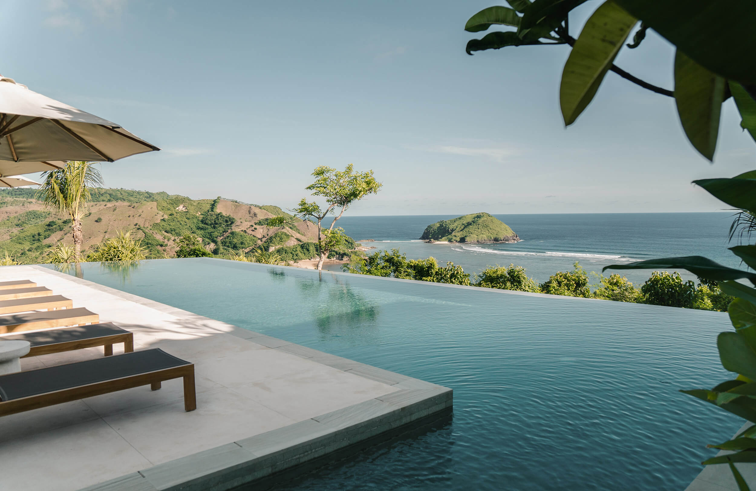 Somewhere Lombok, Indonesia — Infinity pool overlooking coastal cliffs and ocean, teak loungers with cream umbrella, tropical vegetation, minimalist architecture.