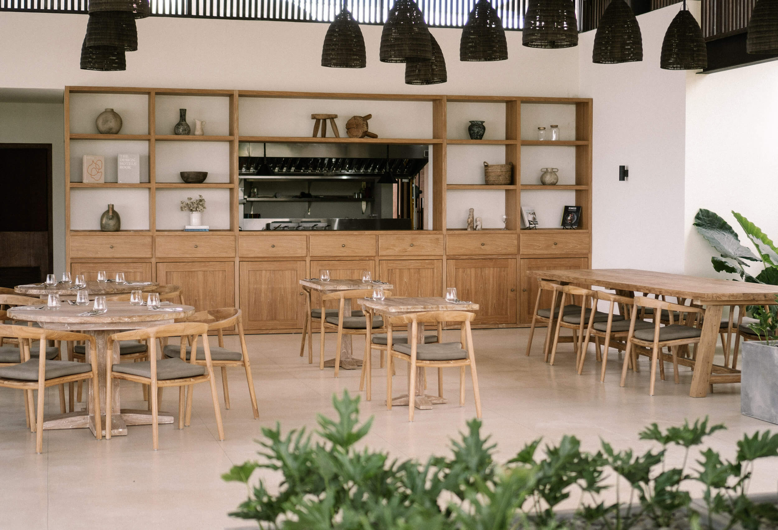 Somewhere Lombok, Indonesia — Modern minimalist dining hall with light wood tables, woven pendant lights, and open kitchen shelving displaying ceramics and glassware.