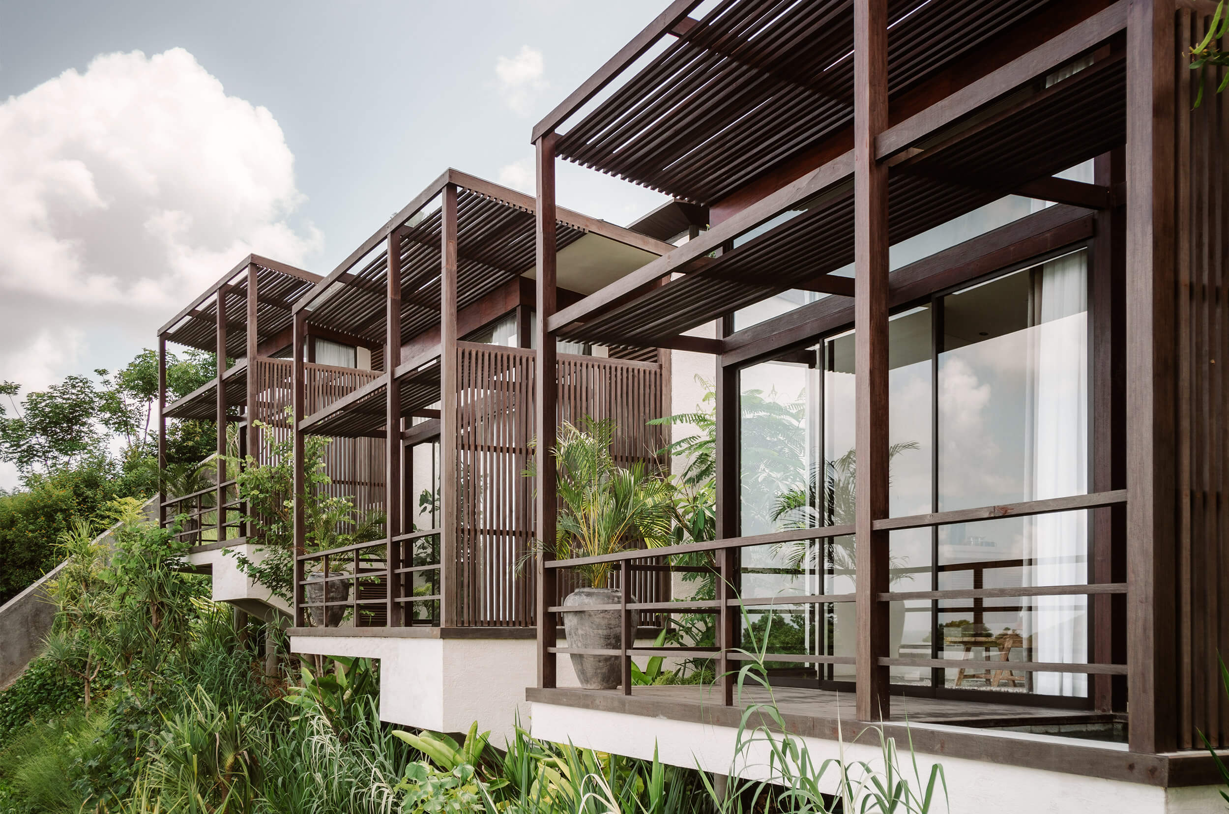 Somewhere Lombok, Asia — Modern timber pavilion with slatted screens, floor-to-ceiling glazing, and tropical garden views under clouded sky.