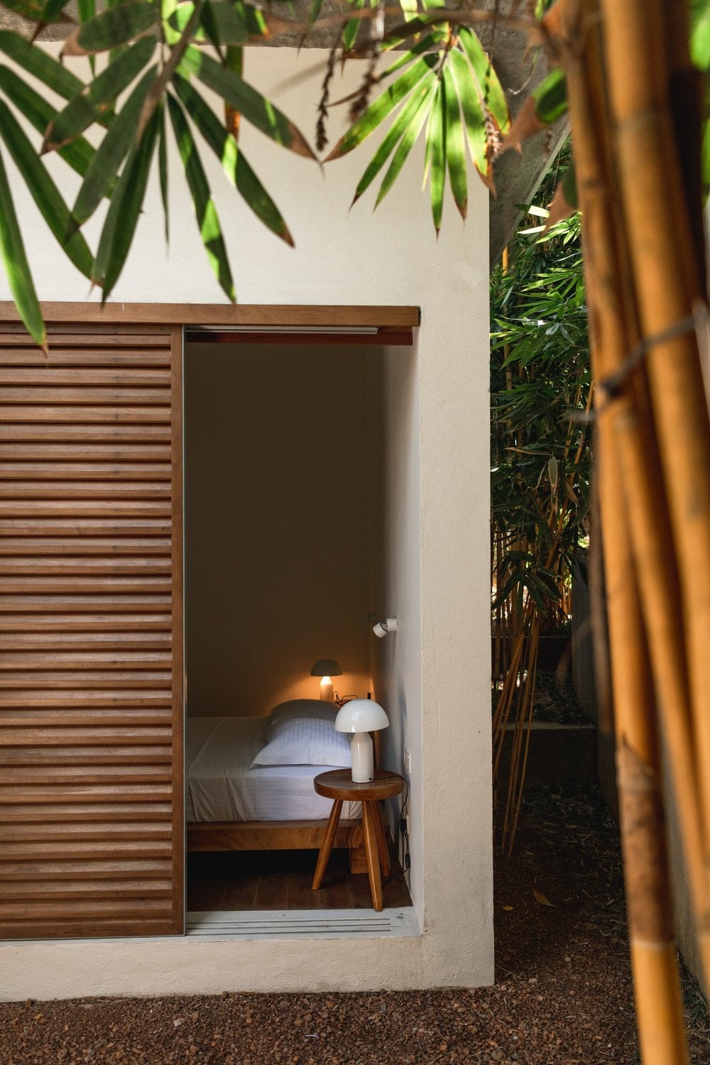 Kurulu Bay, Asia — Minimalist bedroom with slatted wooden door, white walls, and lush bamboo framing in tropical courtyard setting.