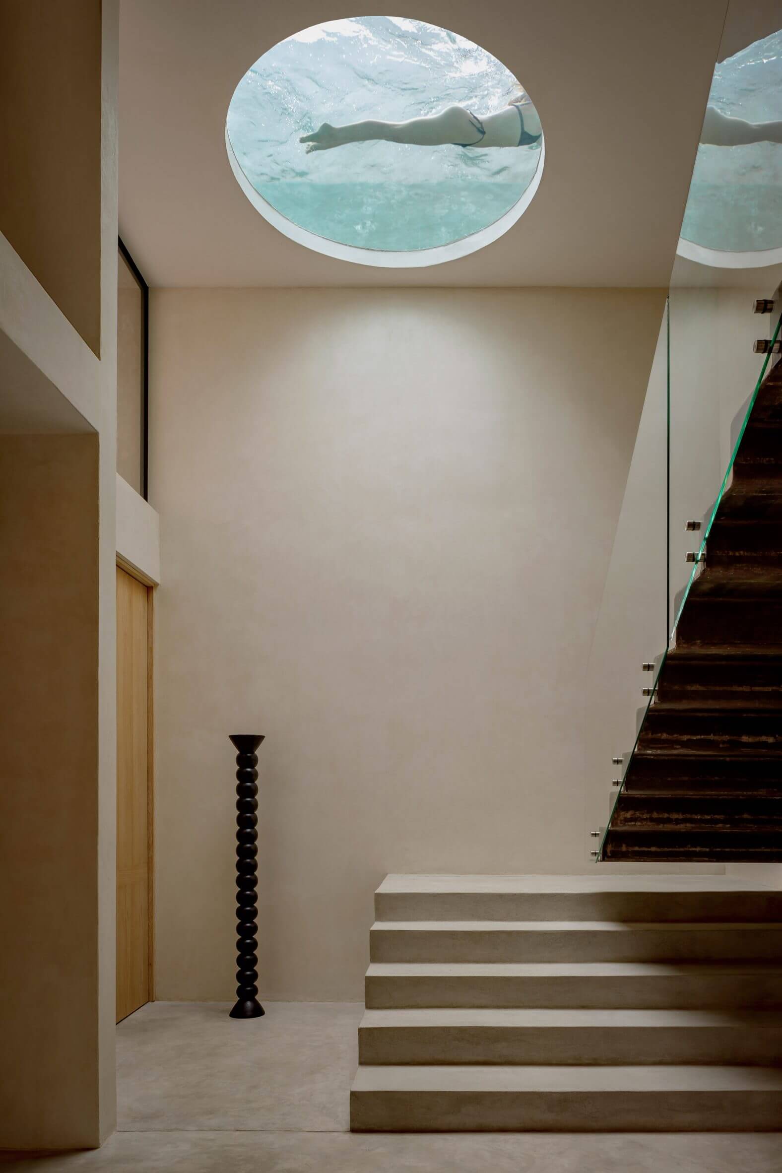 Villa Cava, Mexico — Minimalist staircase with skylight framing swimmer overhead, concrete steps, black sculptural pedestal, warm neutral walls.