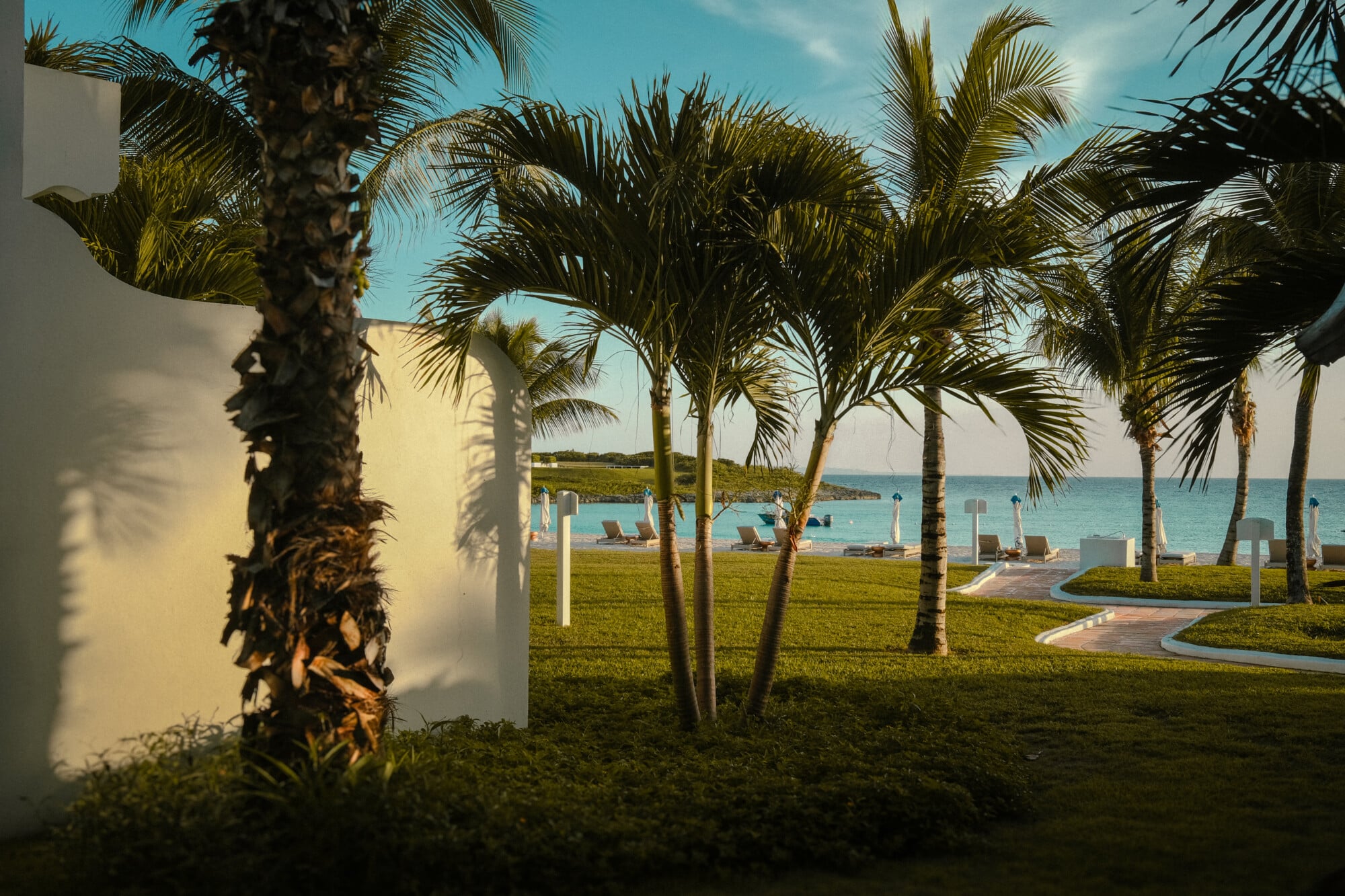 Cap Juluca, Anguilla — Beachfront lawn with coconut palms, white architecture, turquoise Caribbean waters, lounge chairs, and manicured grounds at sunset.