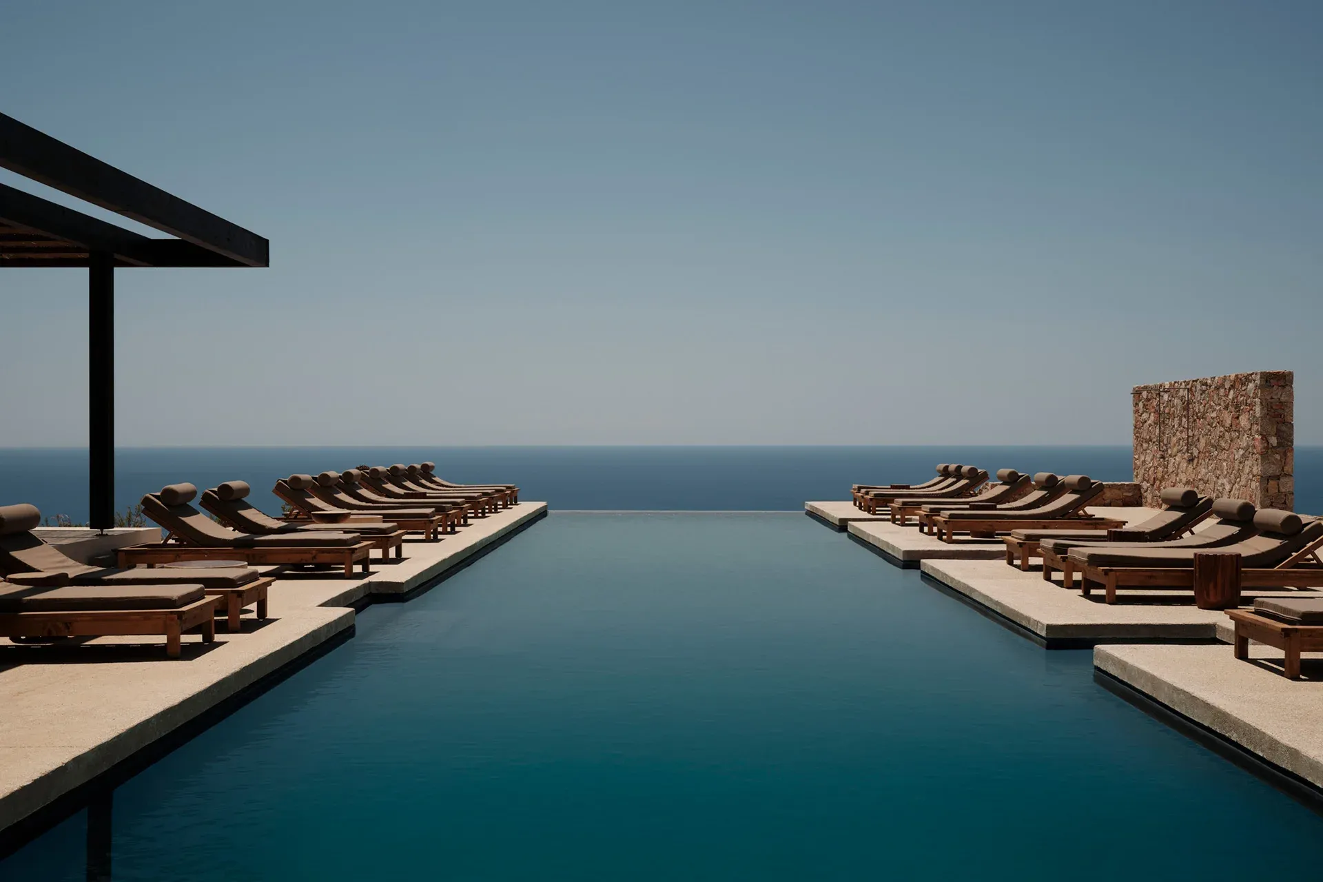 Gundari, Greece — Infinity pool with wooden loungers, stone architecture, and Aegean Sea horizon beneath clear sky.