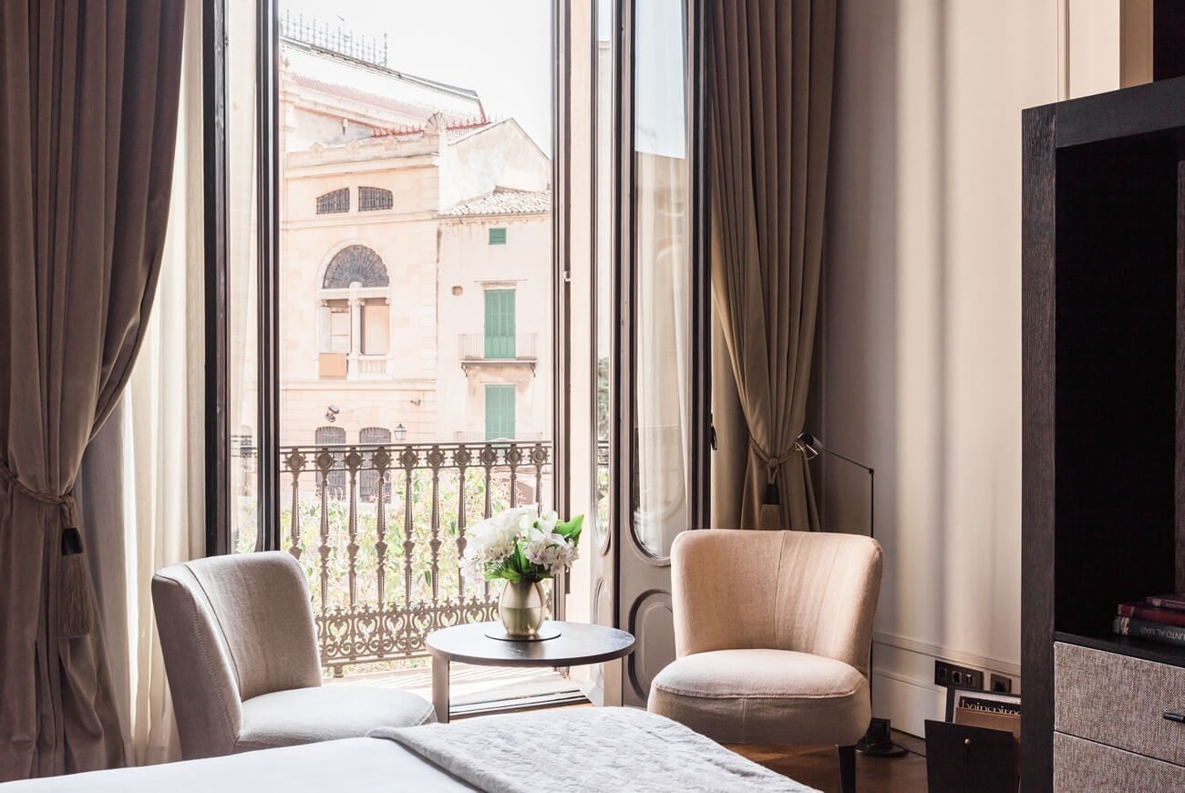 Sant Francesc Hotel Singular, Balearic Islands, Spain — Sunlit hotel room with cream armchairs, ornate wrought-iron balcony overlooking terracotta Mediterranean architecture, flowing taupe curtains.