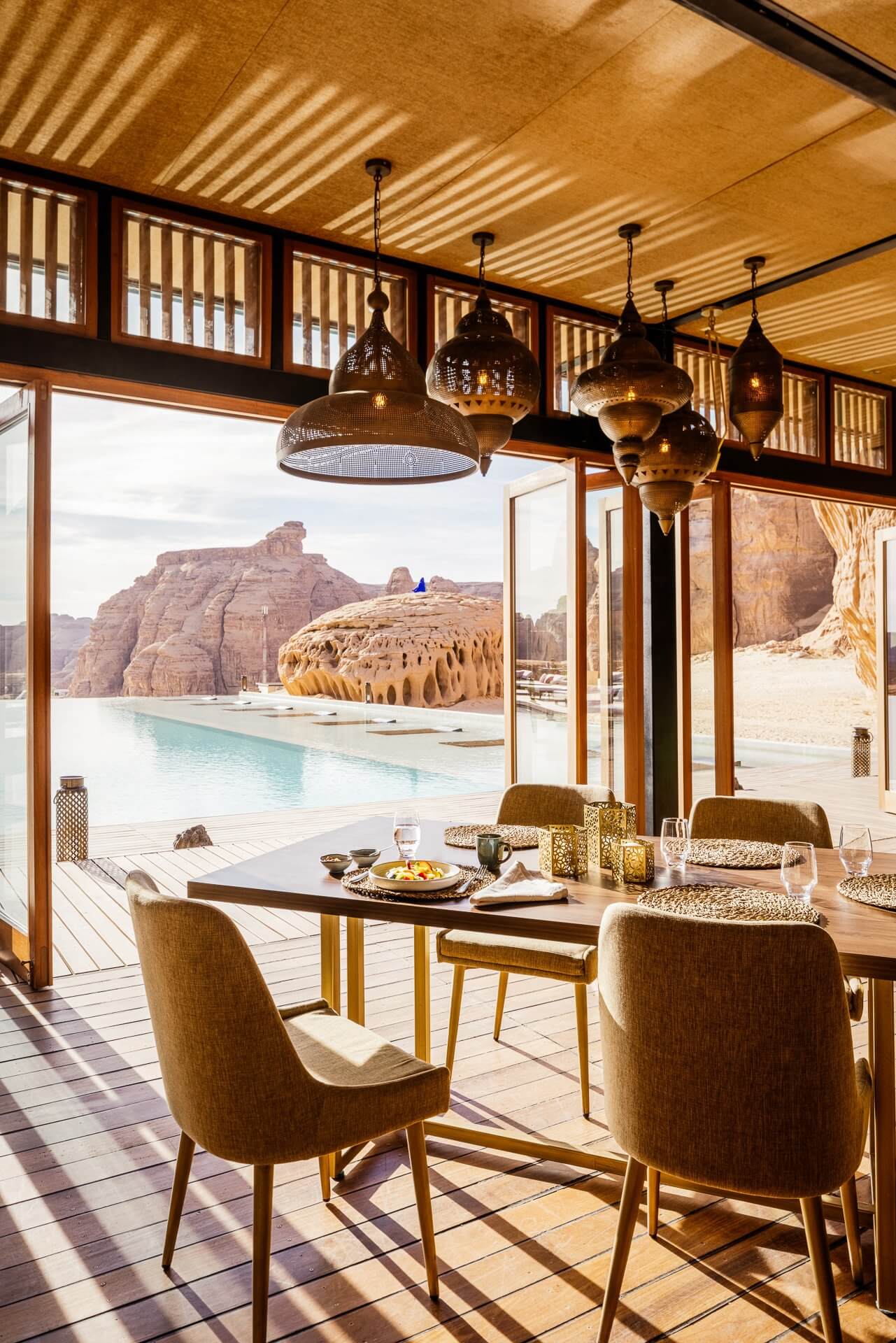 Habitas AlUla, Saudi Arabia — Desert dining pavilion with brass pendant lights, wooden lattice ceiling, turquoise pool and sandstone mountains beyond floor-to-ceiling windows.