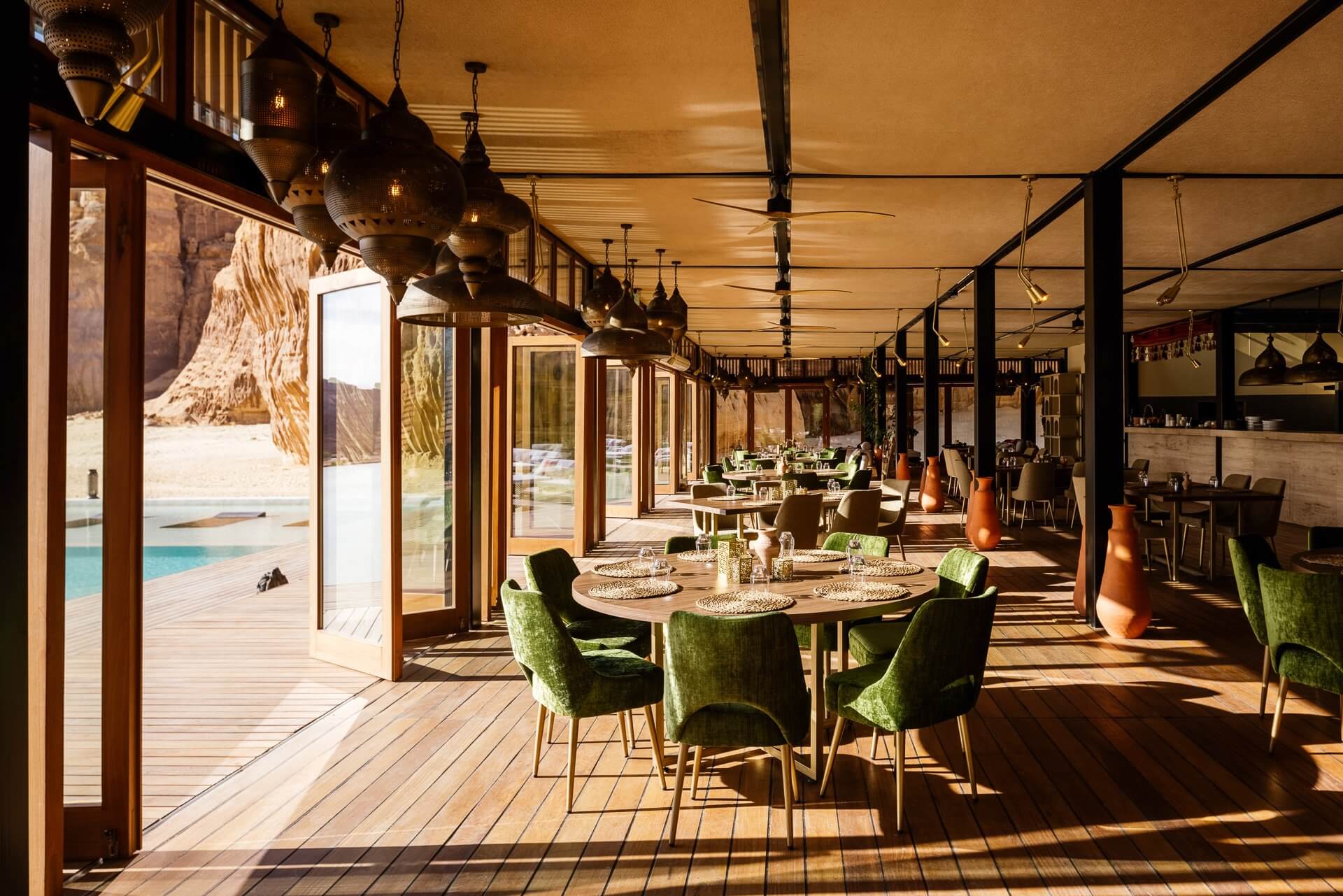 Habitas AlUla — Open-air dining pavilion with brass pendant lights, green upholstered chairs, wooden tables, terracotta vessels, and panoramic desert landscape views.