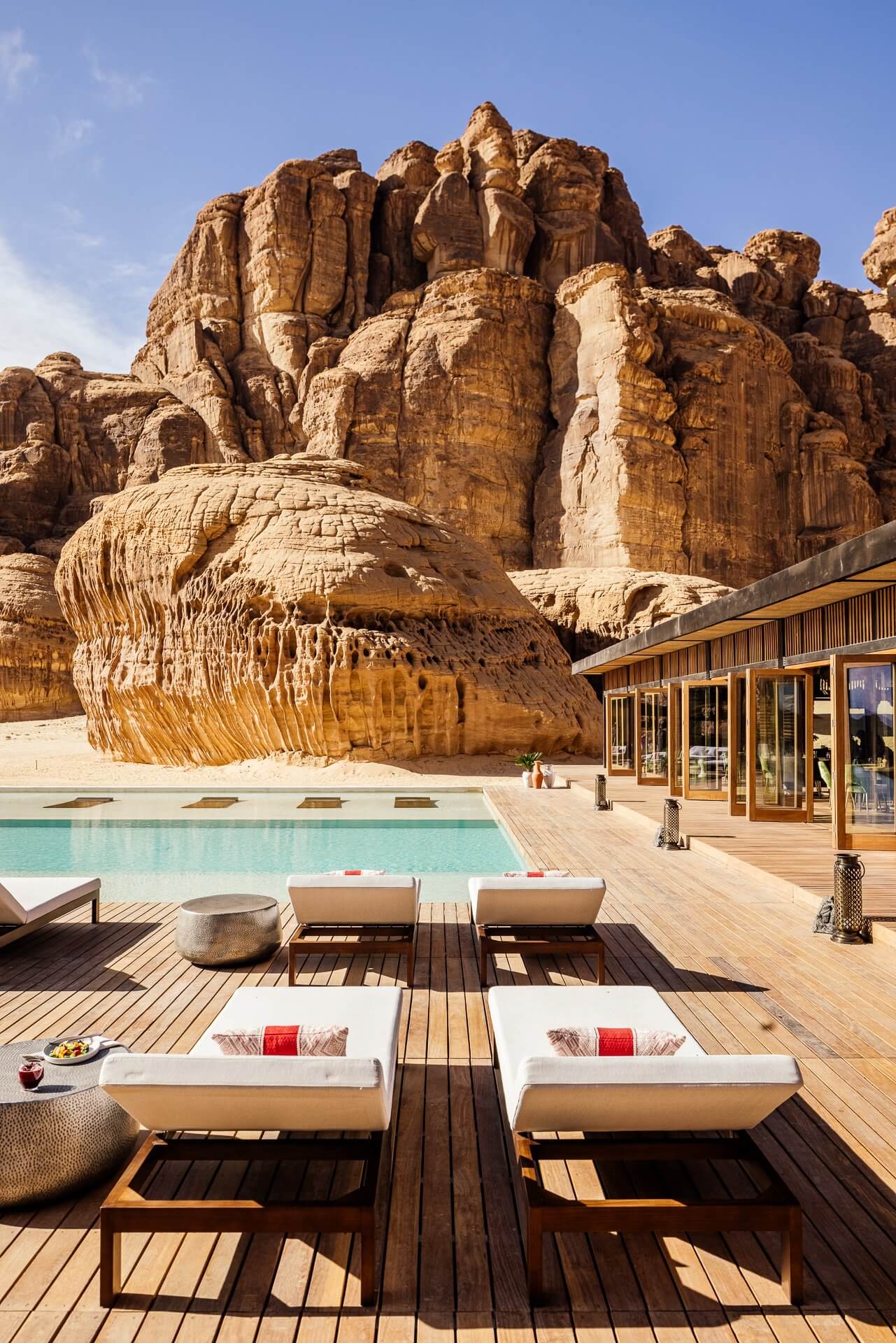 Habitas AlUla, Saudi Arabia — Wooden deck with loungers and turquoise pool overlooking sculptural sandstone rock formations under clear desert sky.