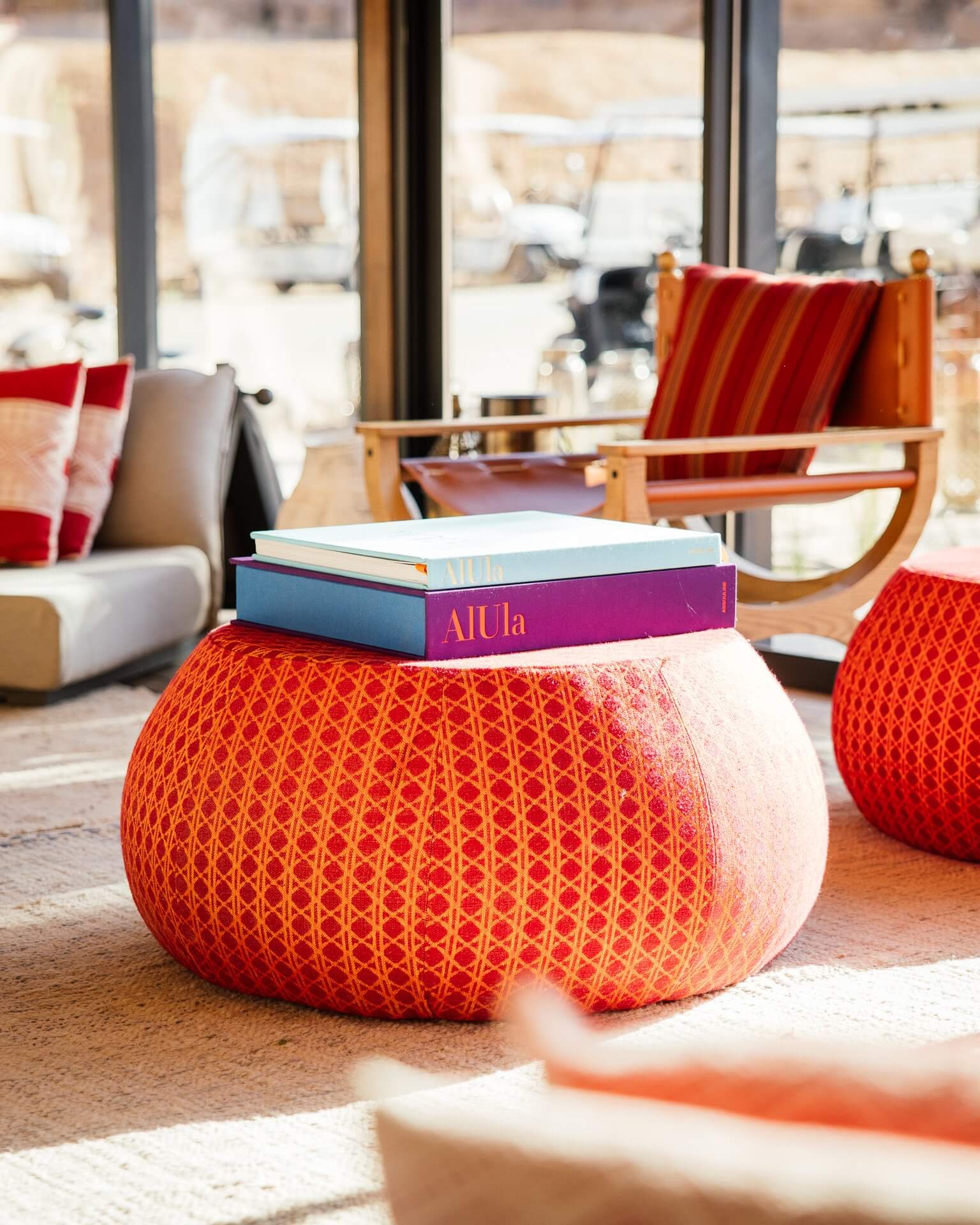Habitas AlUla, Saudi Arabia — Red patterned poufs with stacked books beside wooden chairs and floor-to-ceiling windows overlooking desert landscape.