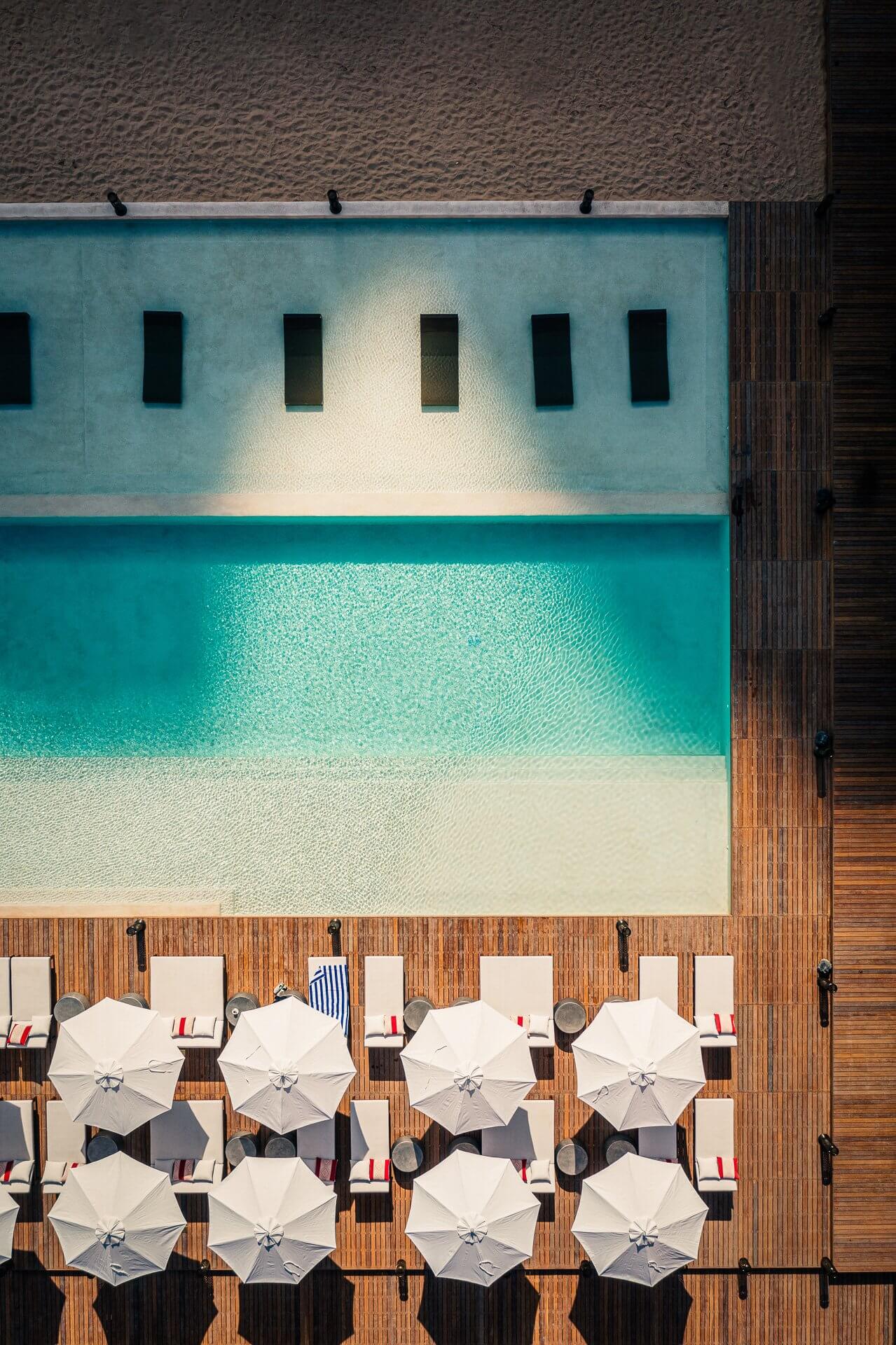 Habitas AlUla — Aerial view of turquoise pool with sandy beach, wooden deck, white umbrellas, and pale blue building with rectangular windows.