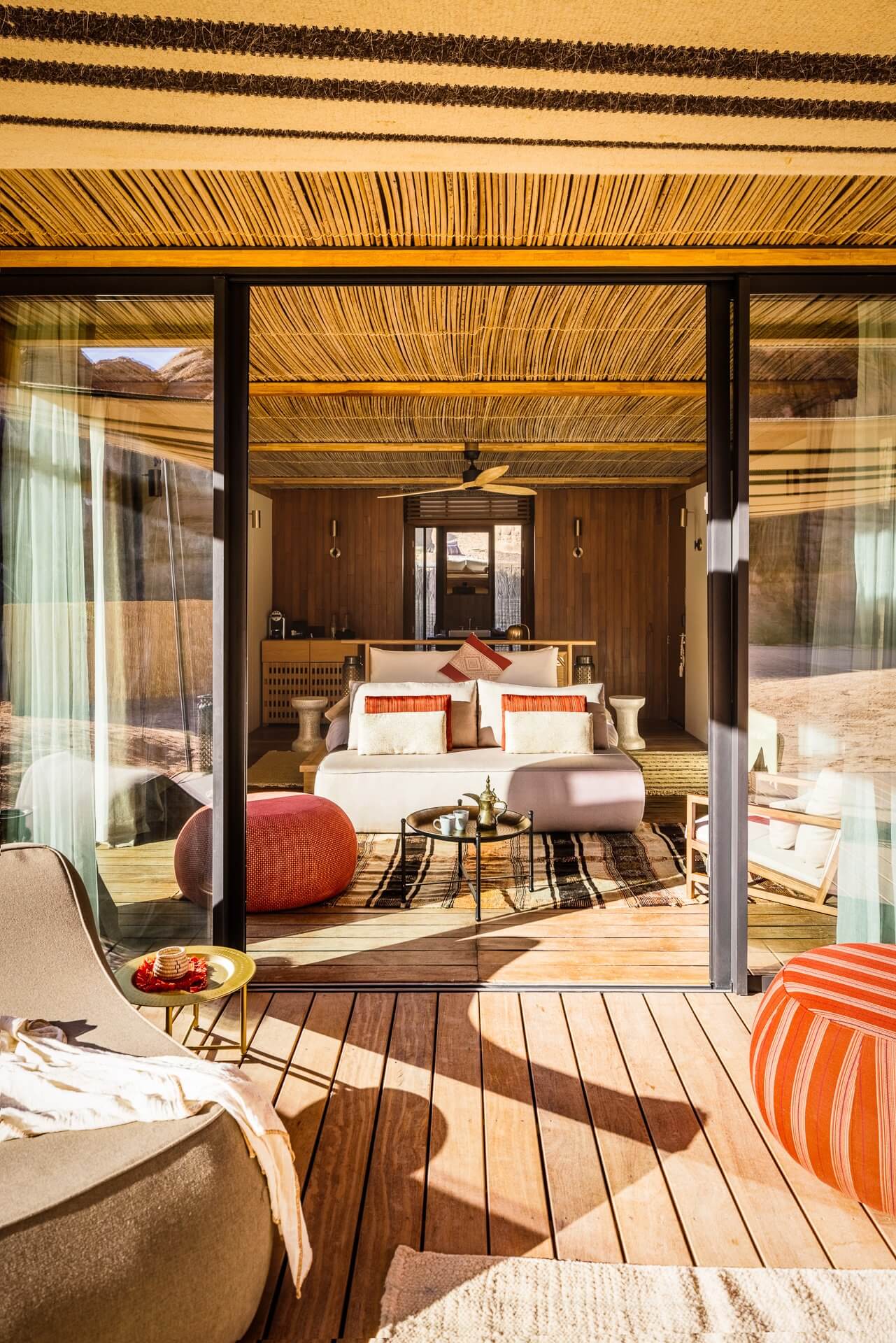 Habitas AlUla — Desert luxury villa with woven ceiling detail, floor-to-ceiling glass doors opening to wooden deck, neutral linens, burnt orange accents, mountain views.