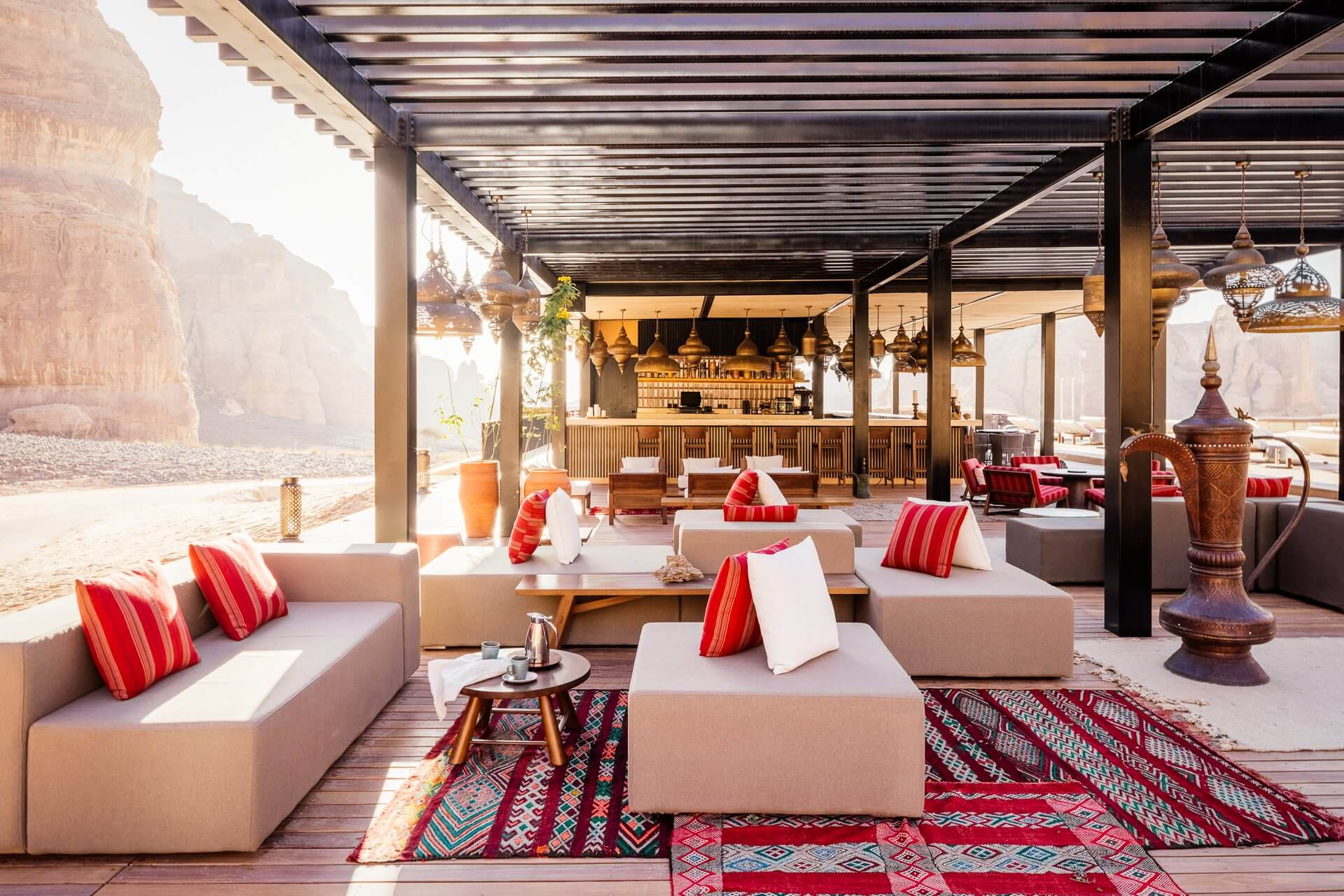Habitas AlUla — Desert resort lounge with cream modular seating, red striped pillows, geometric rugs, brass pendant lights, and sandstone cliffs visible beyond open colonnade.