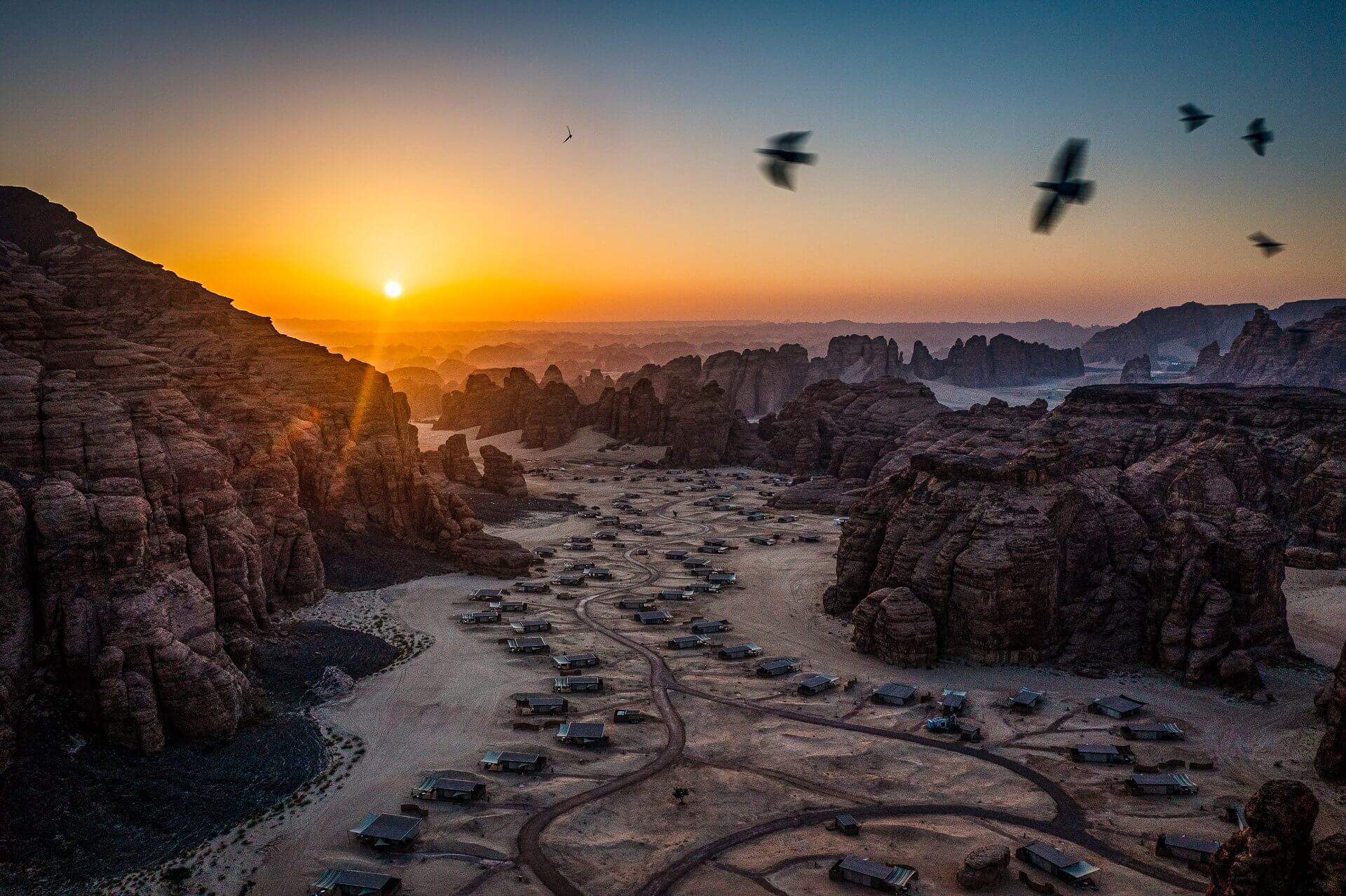 Habitas AlUla, Saudi Arabia — Desert village nestled among towering sandstone formations at sunrise, with traditional dwellings scattered across sandy terrain and birds flying overhead.
