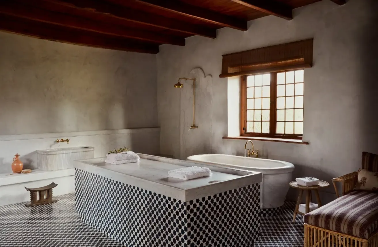 The Bath House — Bath House, Africa — Spa bathroom with black-and-white geometric tile floor, brass fixtures, wooden beams, freestanding tub, and natural light from gridded windows.