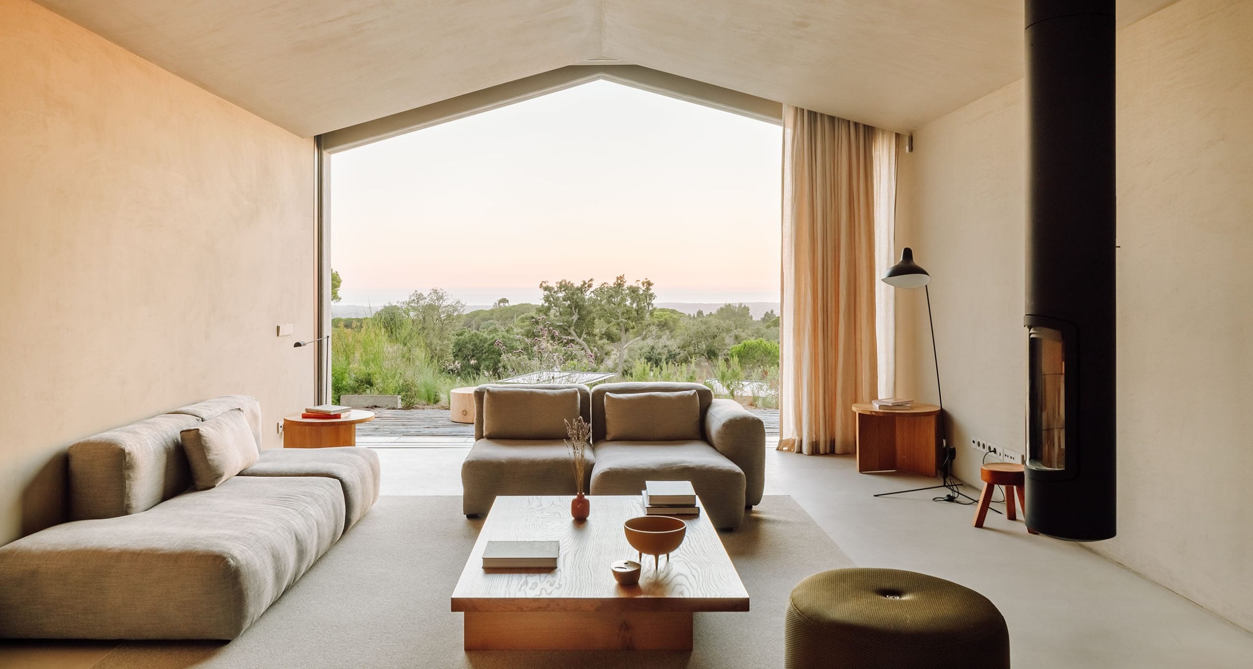 Pa.te.os, Europe — Minimalist living room with curved cream sofas, wooden coffee table, and expansive windows overlooking Mediterranean landscape at dusk.