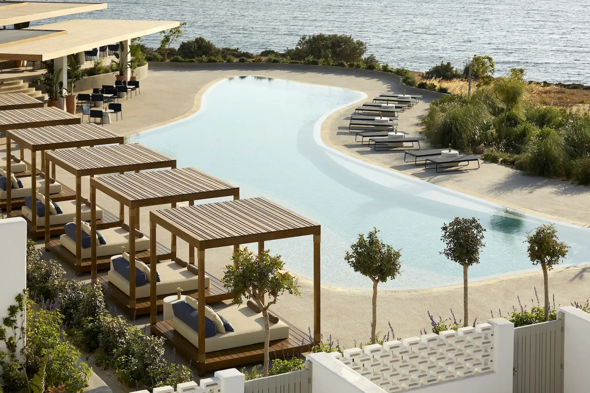 Pārocks, Europe — Curved luxury pool with pale stone surround, wooden cabanas, manicured landscaping, and calm water views at dusk.