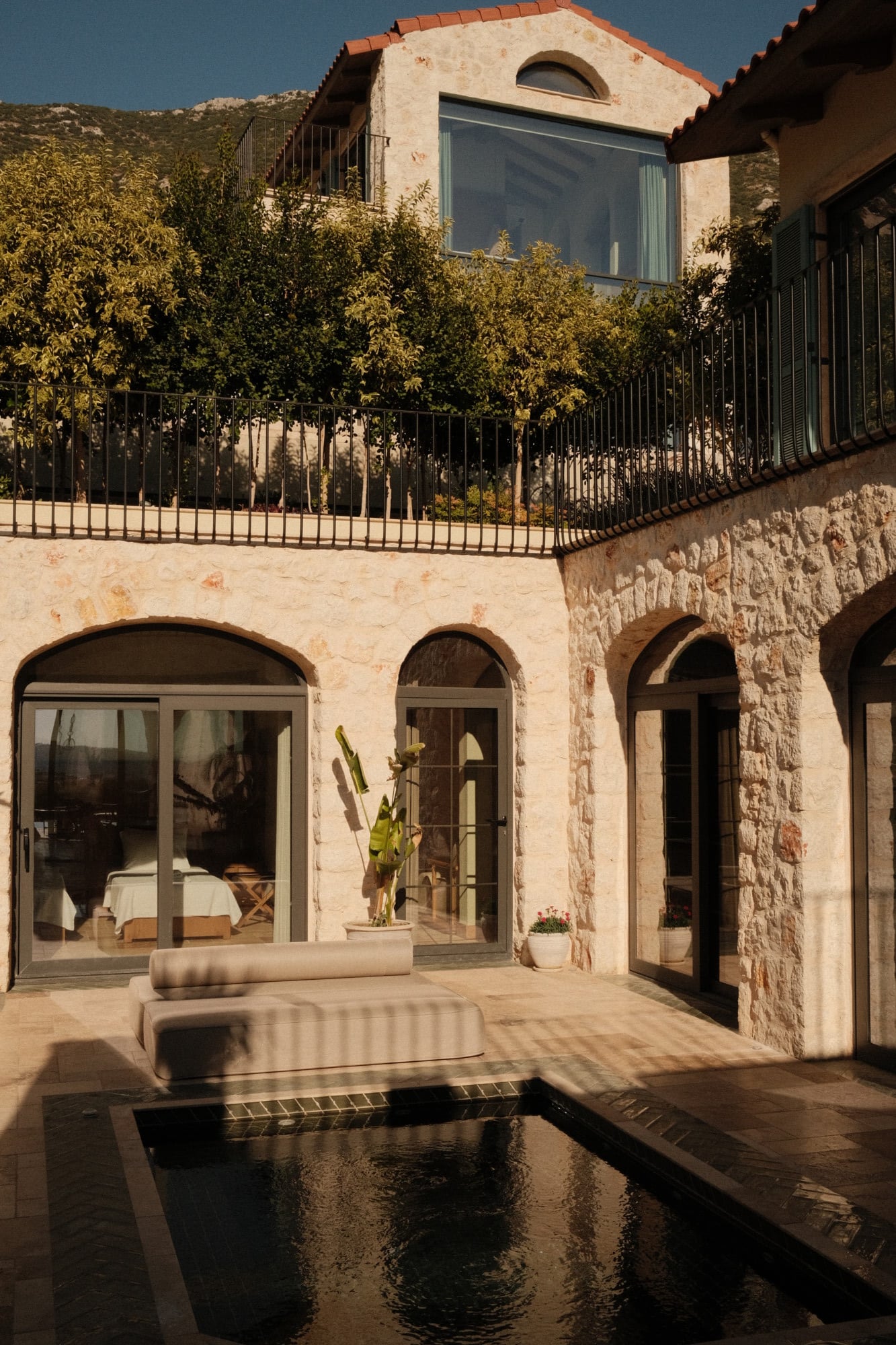 Villa Thalia by Muses Kalkan, Kalkan, Turkey — Minimalist courtyard with arched stone colonnade, reflecting pool, contemporary furniture, and olive tree-covered hillside backdrop.