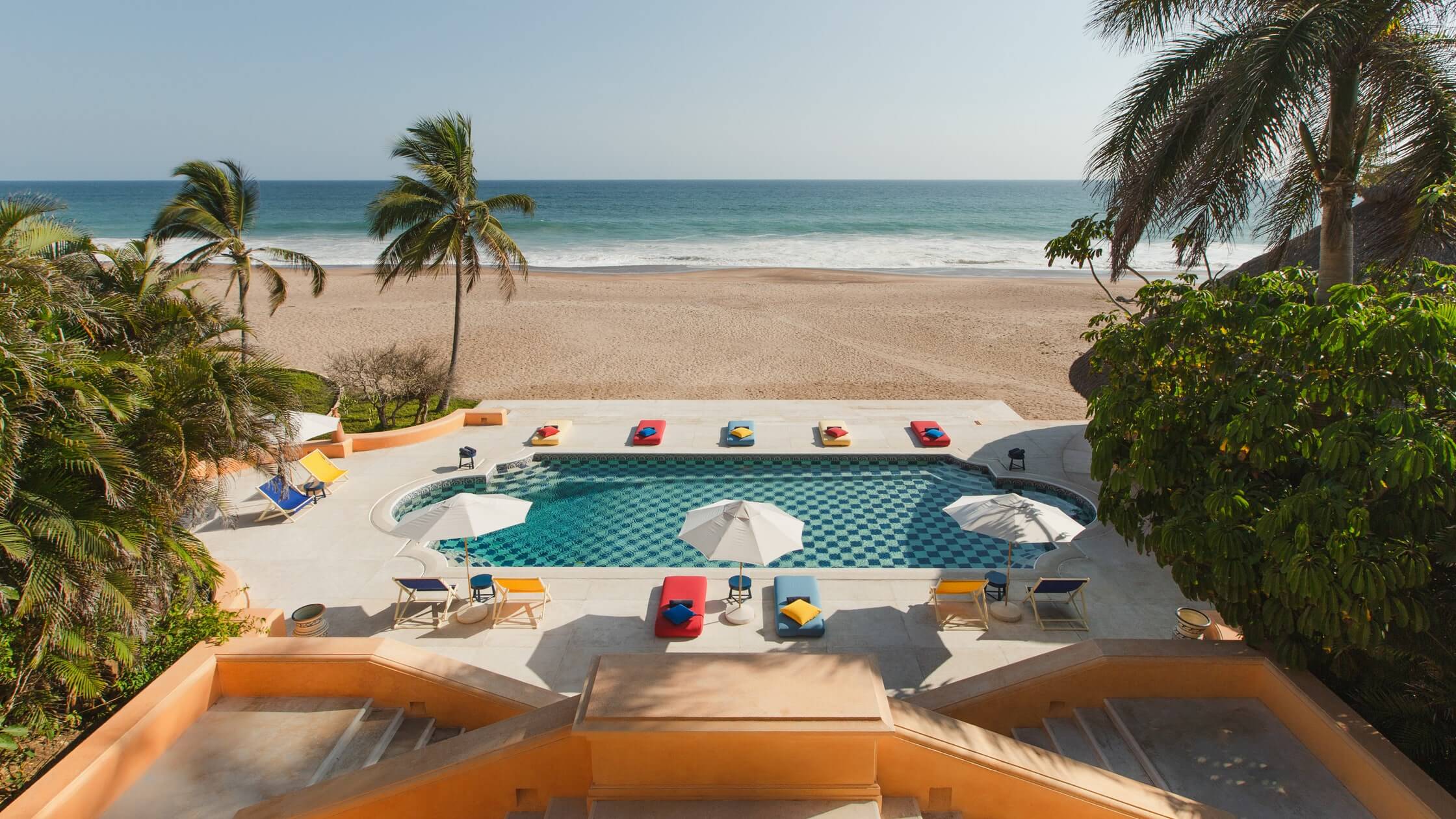 Cuixmala, Mexico — Oceanfront pool with turquoise tile, white umbrellas, and colorful loungers overlooking pristine beach and palm trees.
