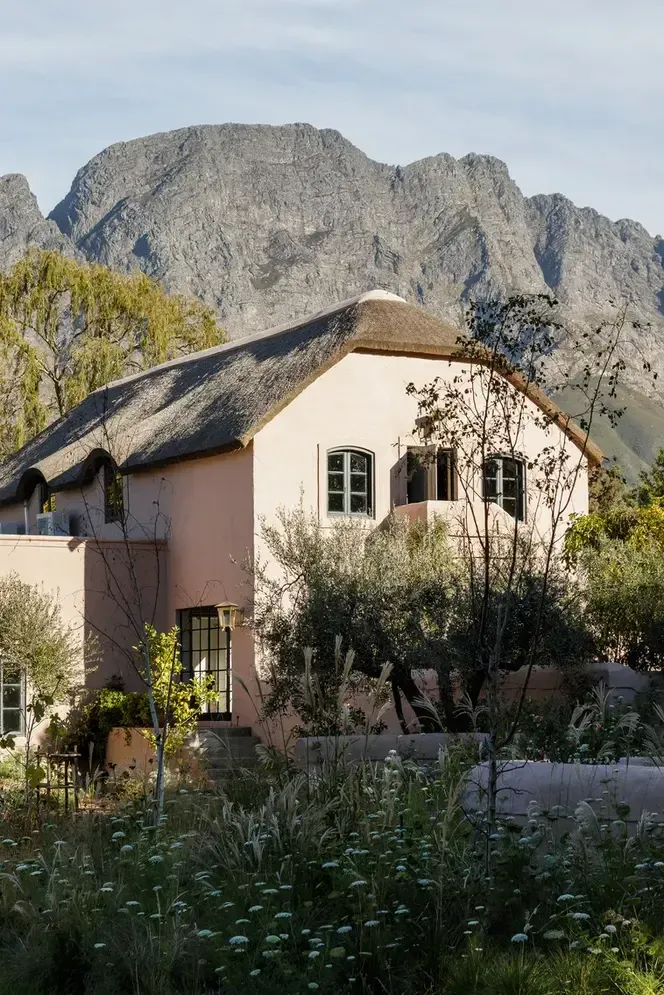 Sterrekopje, Africa — Cream-rendered cottage with thatched roof beneath dramatic rocky mountain, surrounded by indigenous plantings and wildflowers.
