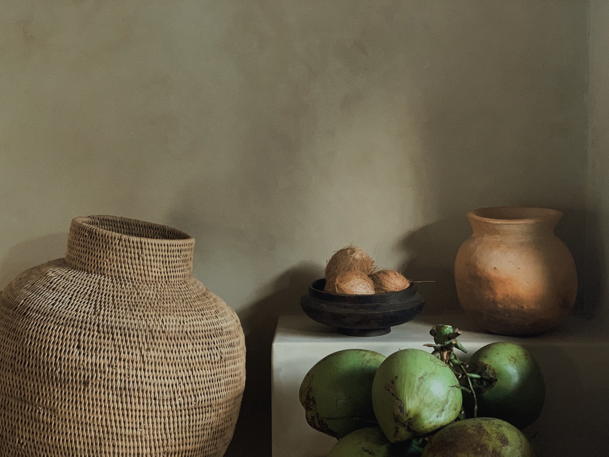 Sussurro, Africa — Woven basket, terracotta vessels, and fresh green fruits arranged on white surface with warm natural light casting shadows against neutral wall.