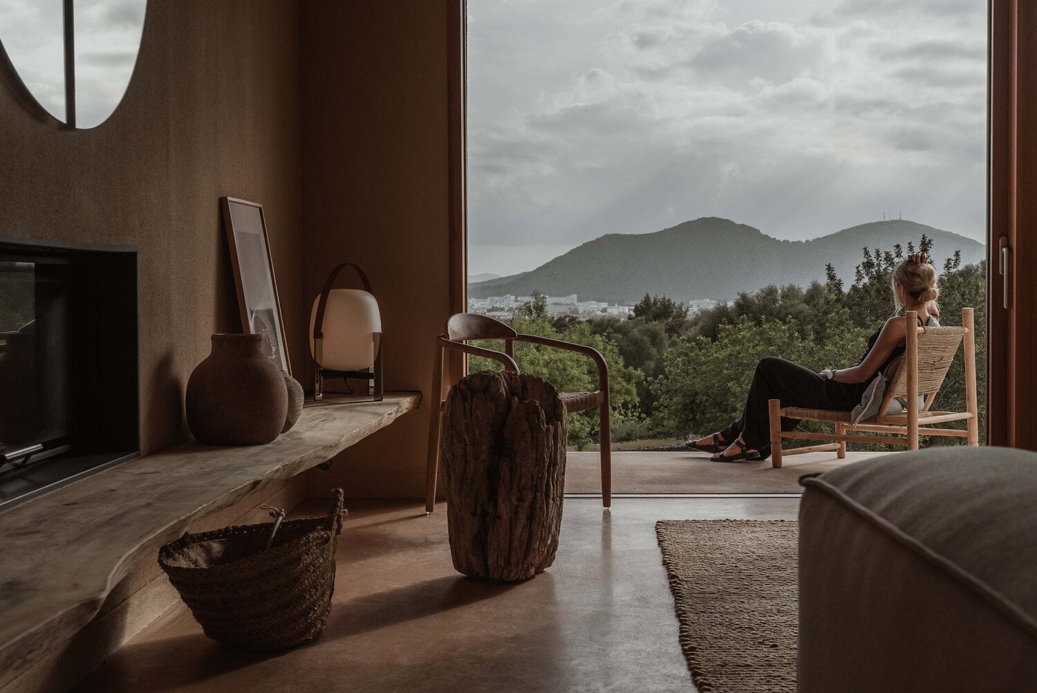 Alcúdia House, Balearic Islands — Woman seated on woven chair overlooking forested mountains, earth-toned interior with concrete floors and natural materials.